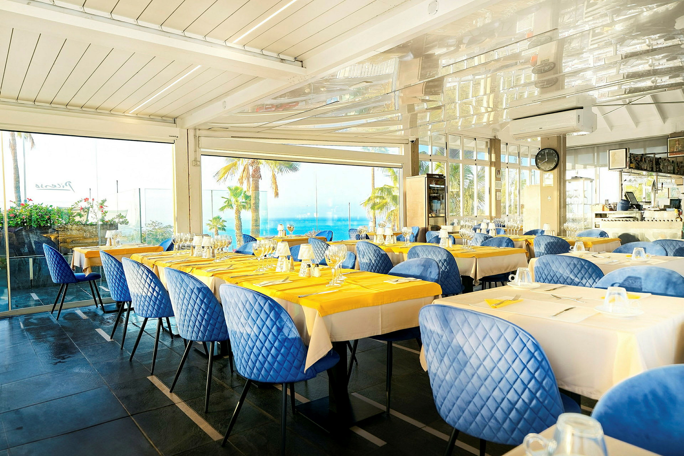 Restaurang med dukade bord, blå stolar och utsikt över palmer och havet genom stora fönster. (AI-genererad text)