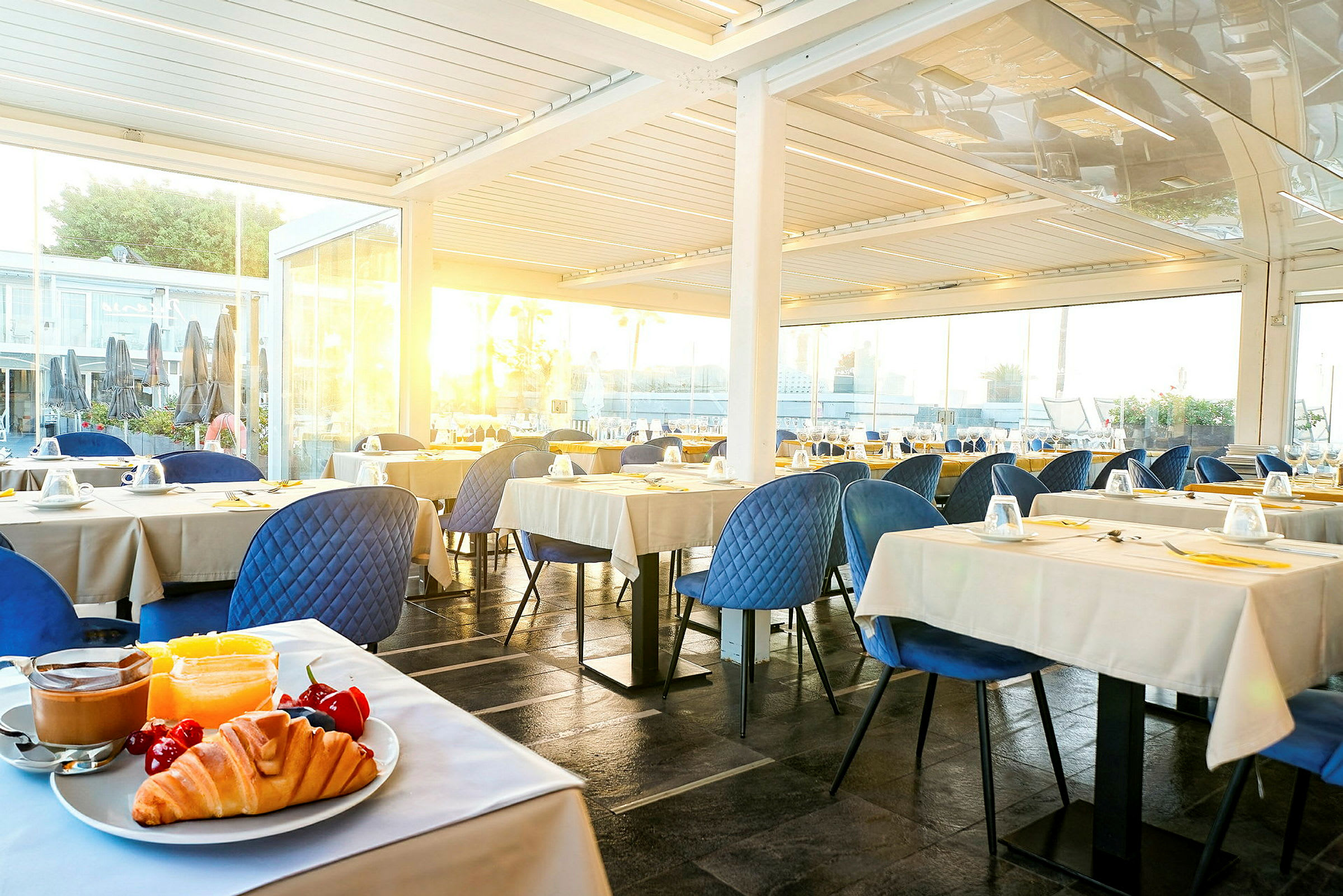 Bild av en restaurang med bordsdukning. I förgrunden syns en tallrik med croissant, frukt och kaffe. (AI-genererad text)
