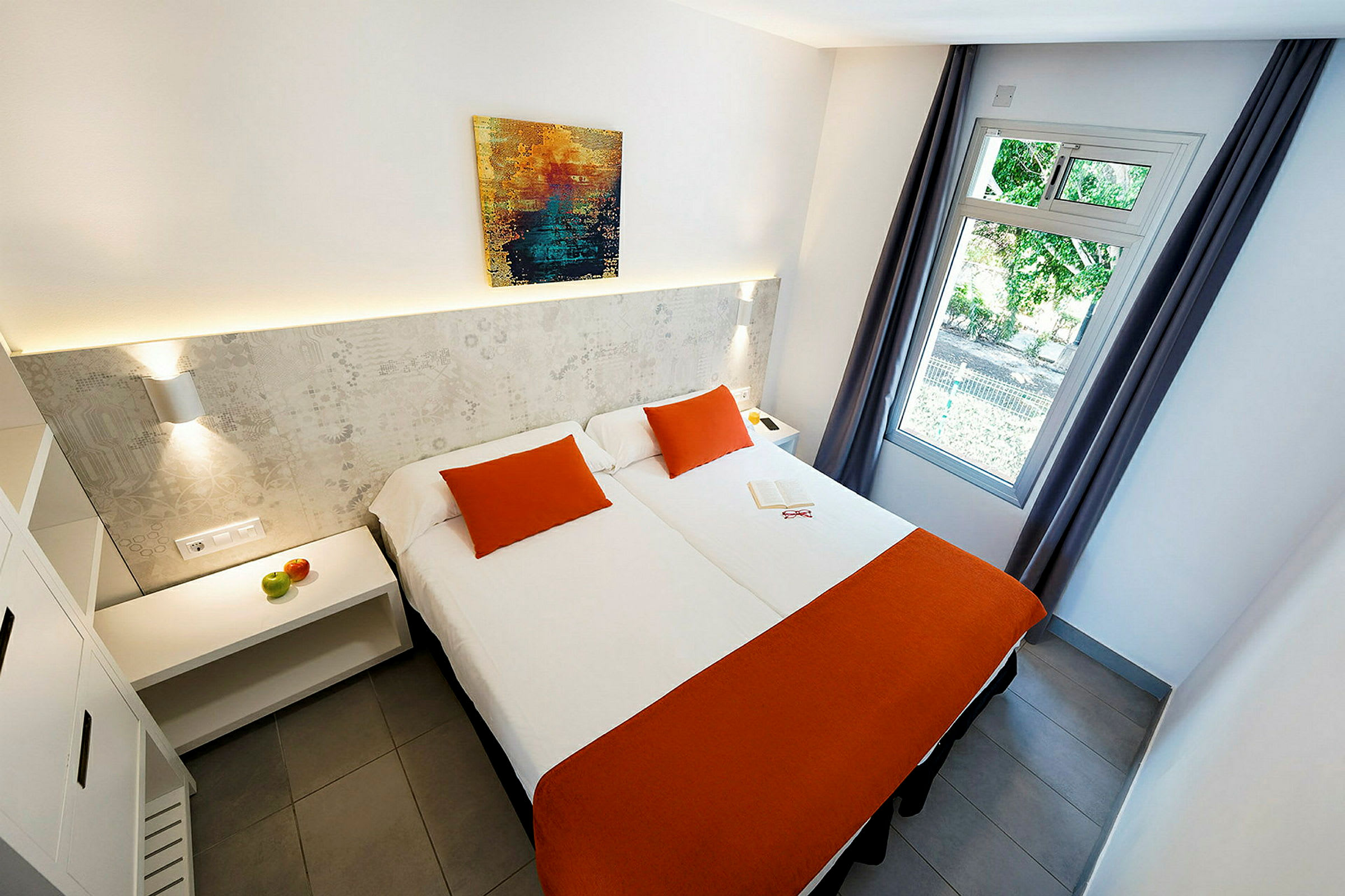 Ett hotellrum med två sängar med orange överkast och prydnadskuddar, en tavla på väggen och ett öppet fönster med sidodraperier. (AI-genererad text)