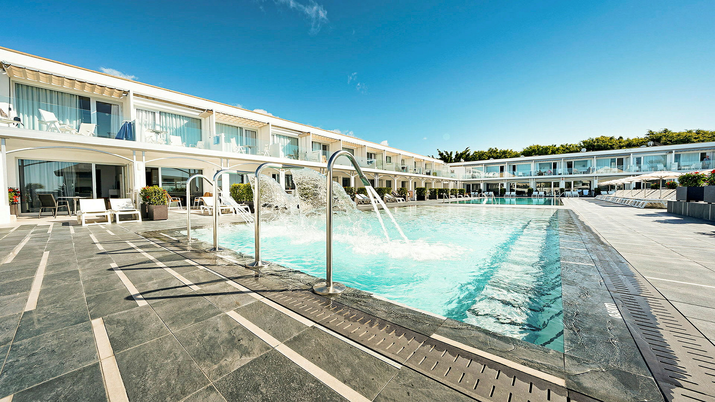 En utomhuspool med splashfunktioner inför en rad hotellrum med uteplatser. (AI-genererad text)