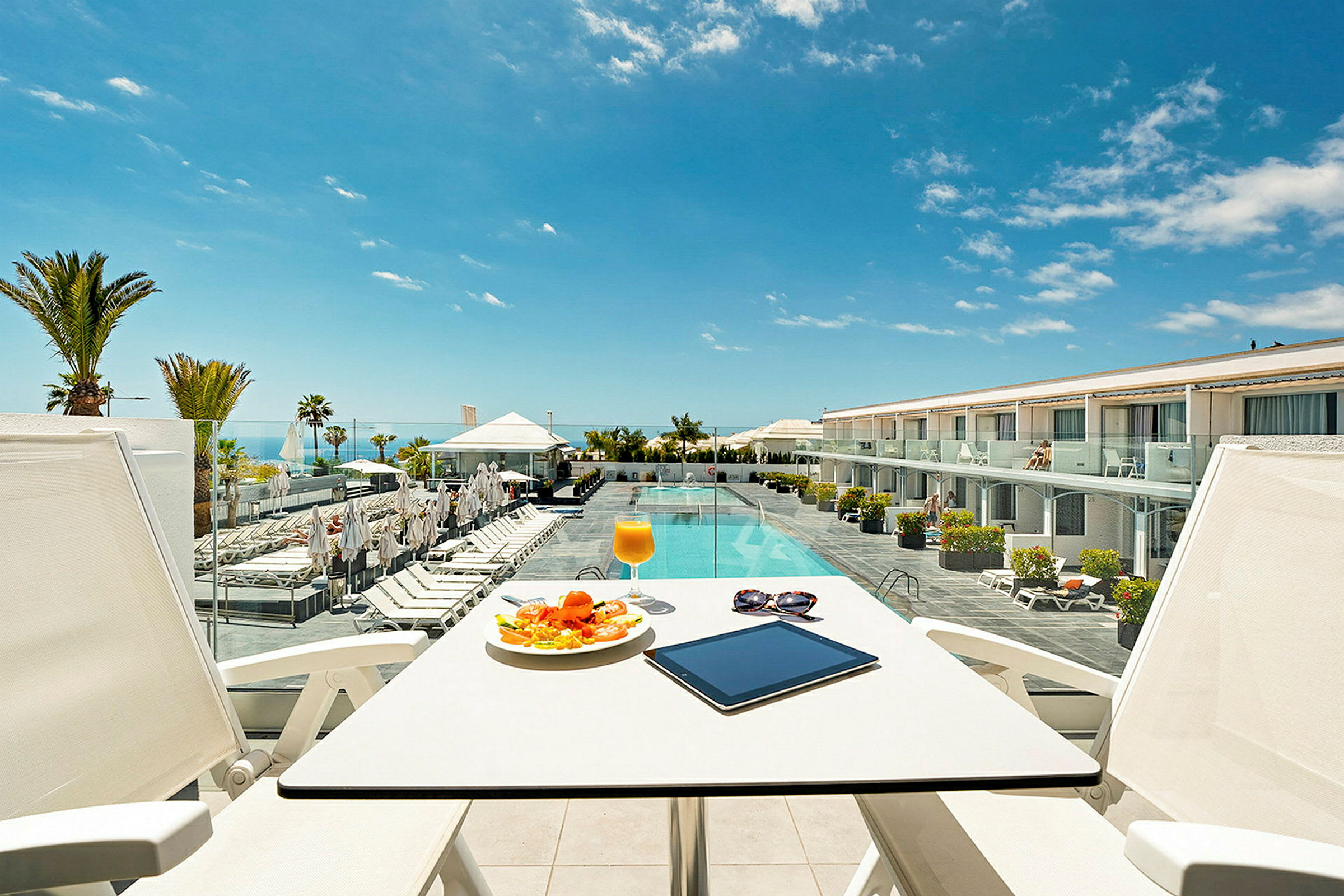 Utsikt från en terrass med bord och stolar mot en pool och hotellbyggnad. (AI-genererad text)