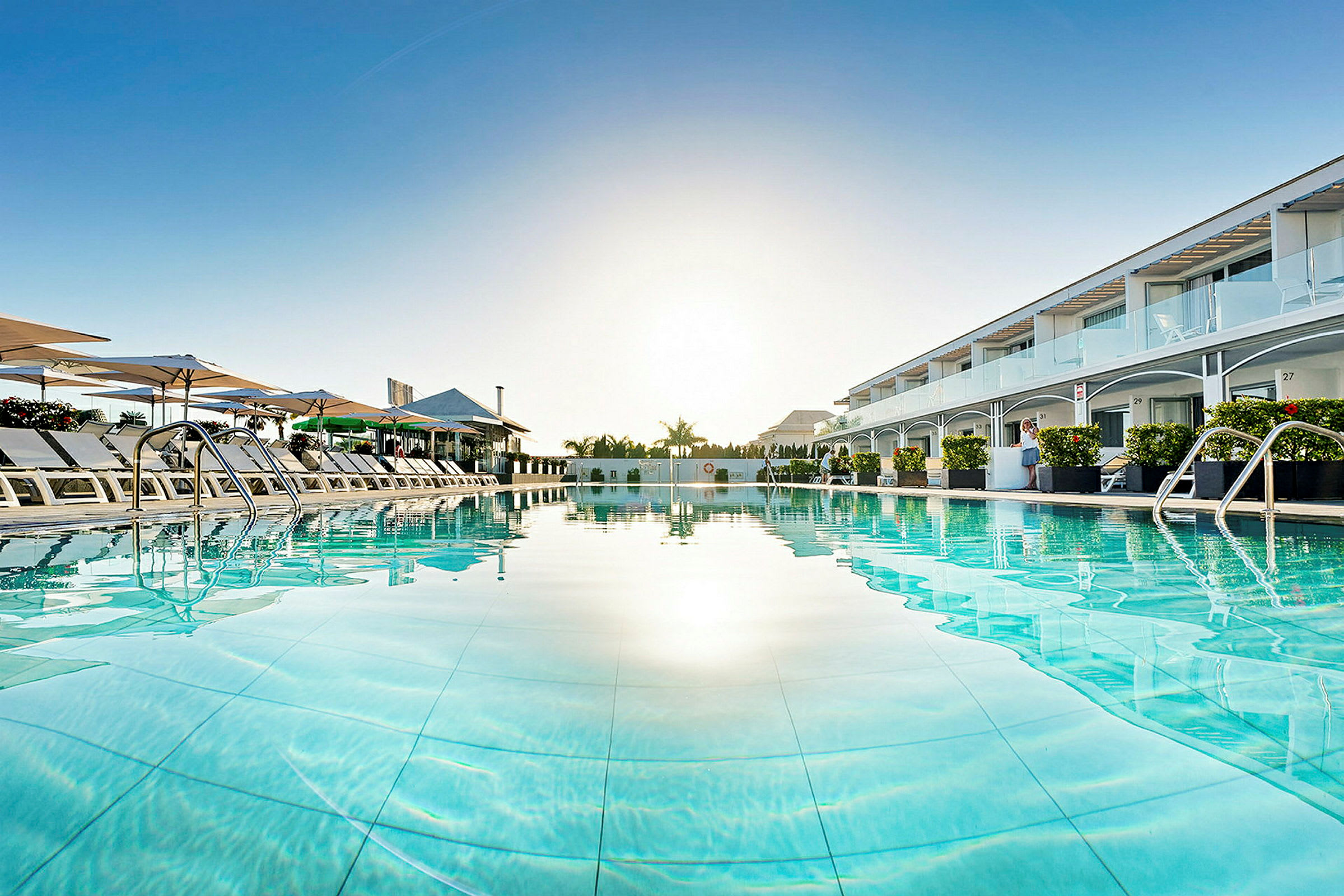 Utsikt över en pool med solstolar och parasoller, omgiven av hotellbyggnader. (AI-genererad text)