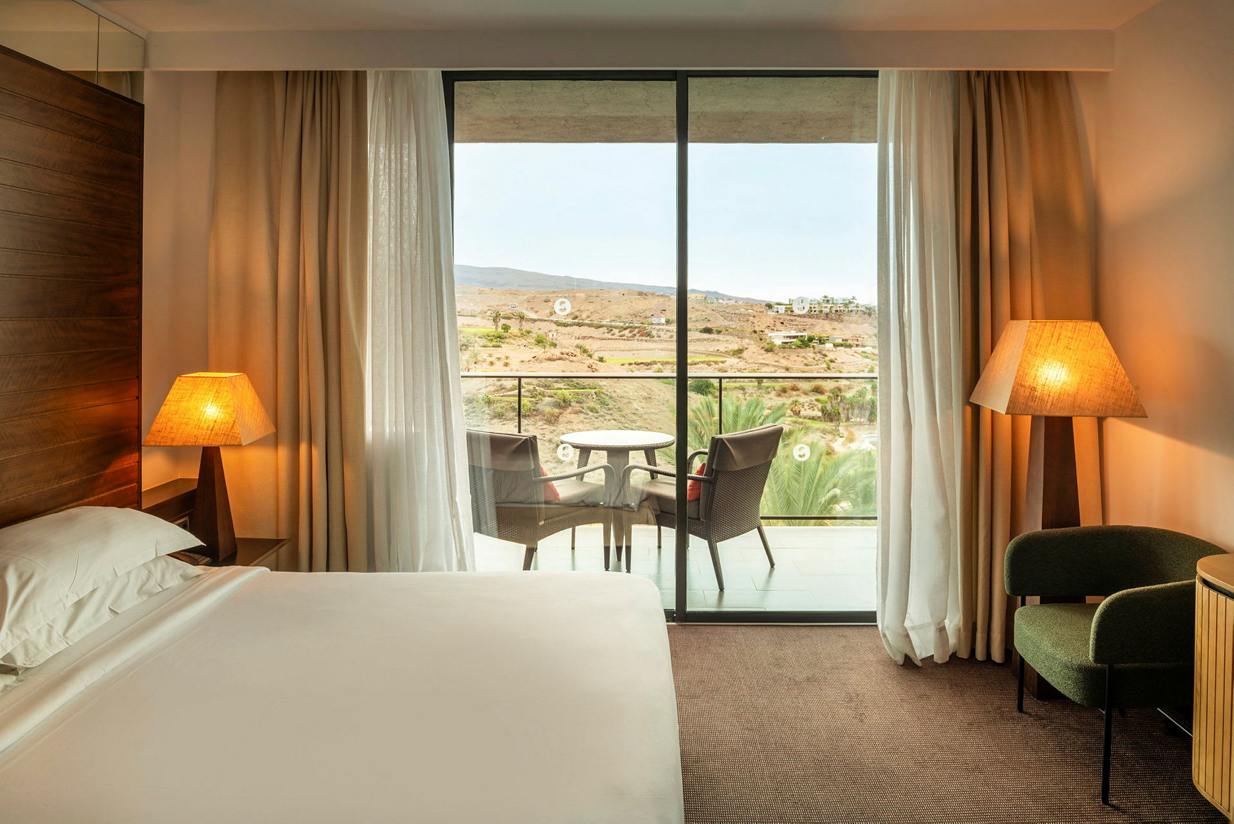 Ett hotellrum med en säng, två bordslampor, en fåtölj och en balkong med sittmöbler. (AI-genererad text)