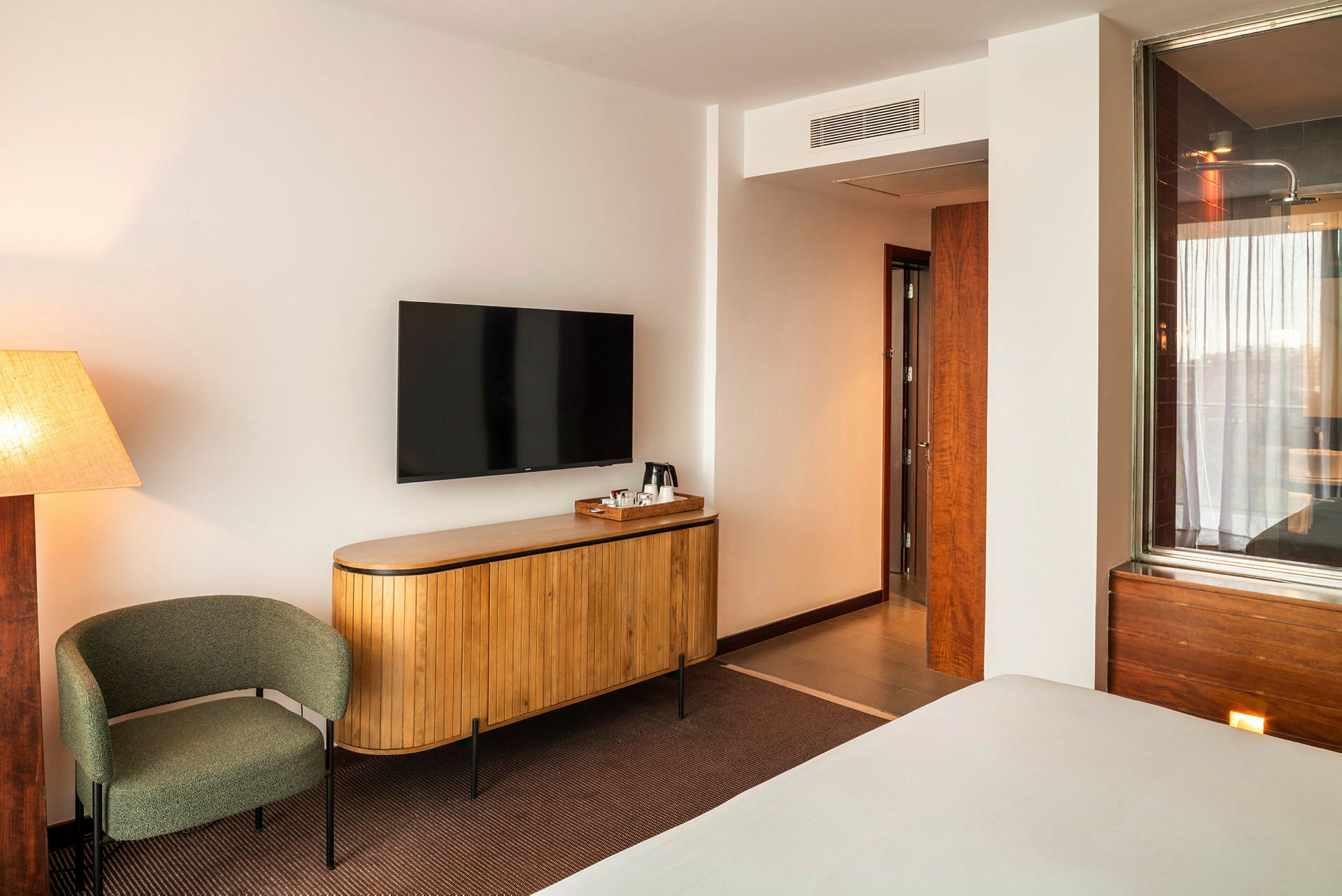 Ett hotellrum med beige väggar, en svängbar stol, en elegant TV monterad på väggen ovanför en träskänk och en dörr som leder till badrummet. (AI-genererad text)