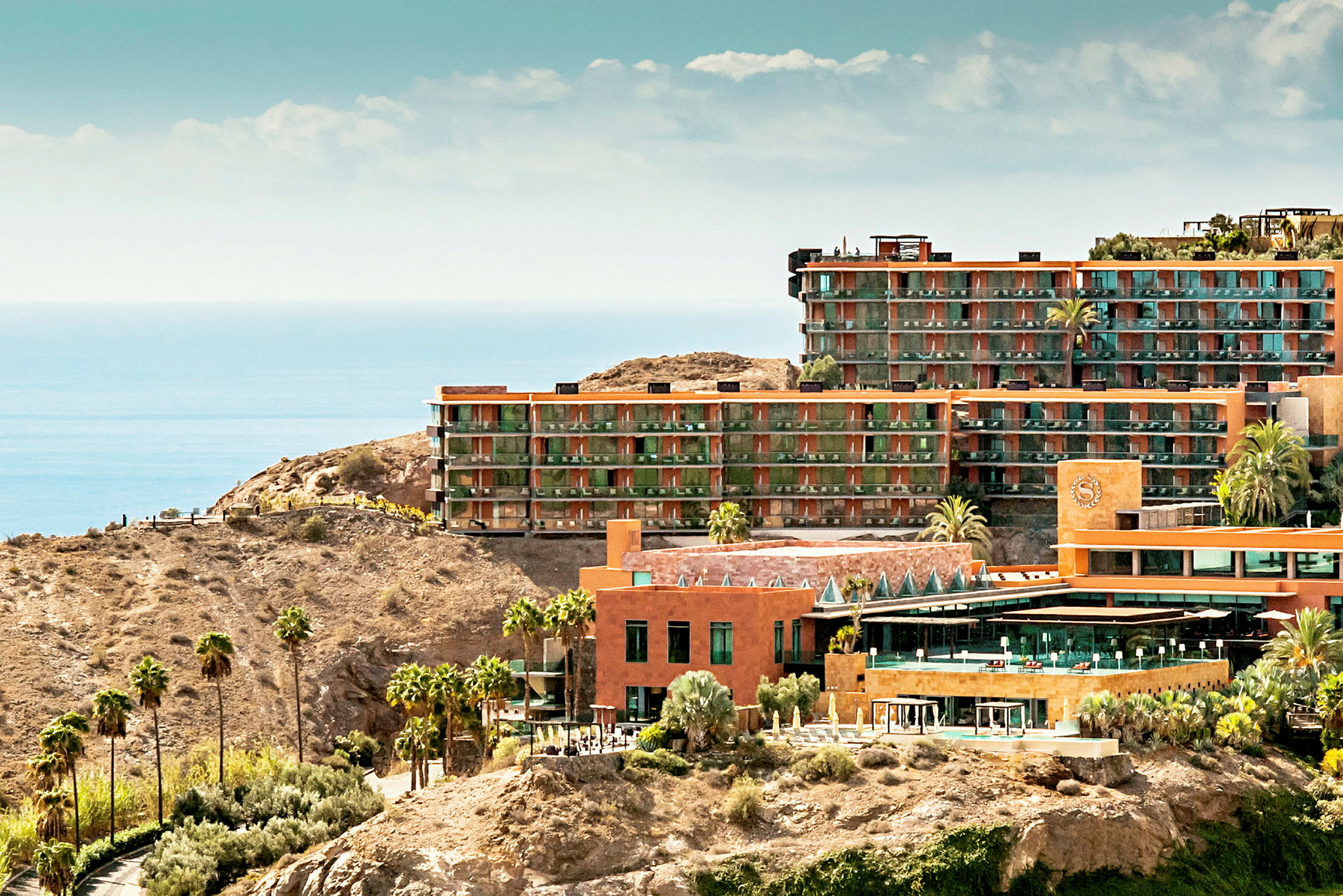 Bild på hotellbyggnader i terrakottastil på klippig terräng med utsikt över havet. (AI-genererad text)