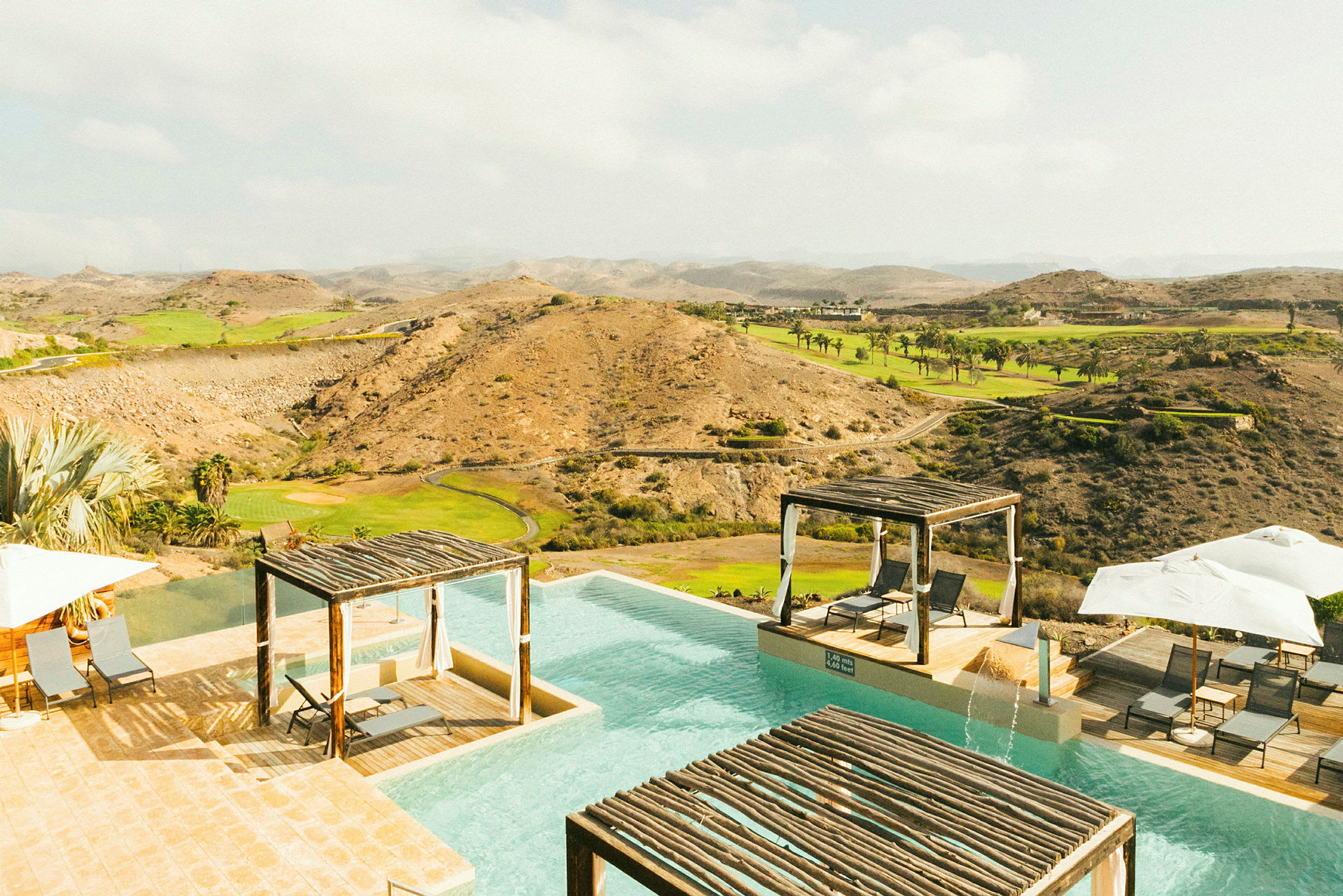 En infinitypool med solstolar och paviljonger vid kanten, omgiven av ett kargt landskap med gröna områden. (AI-genererad text)