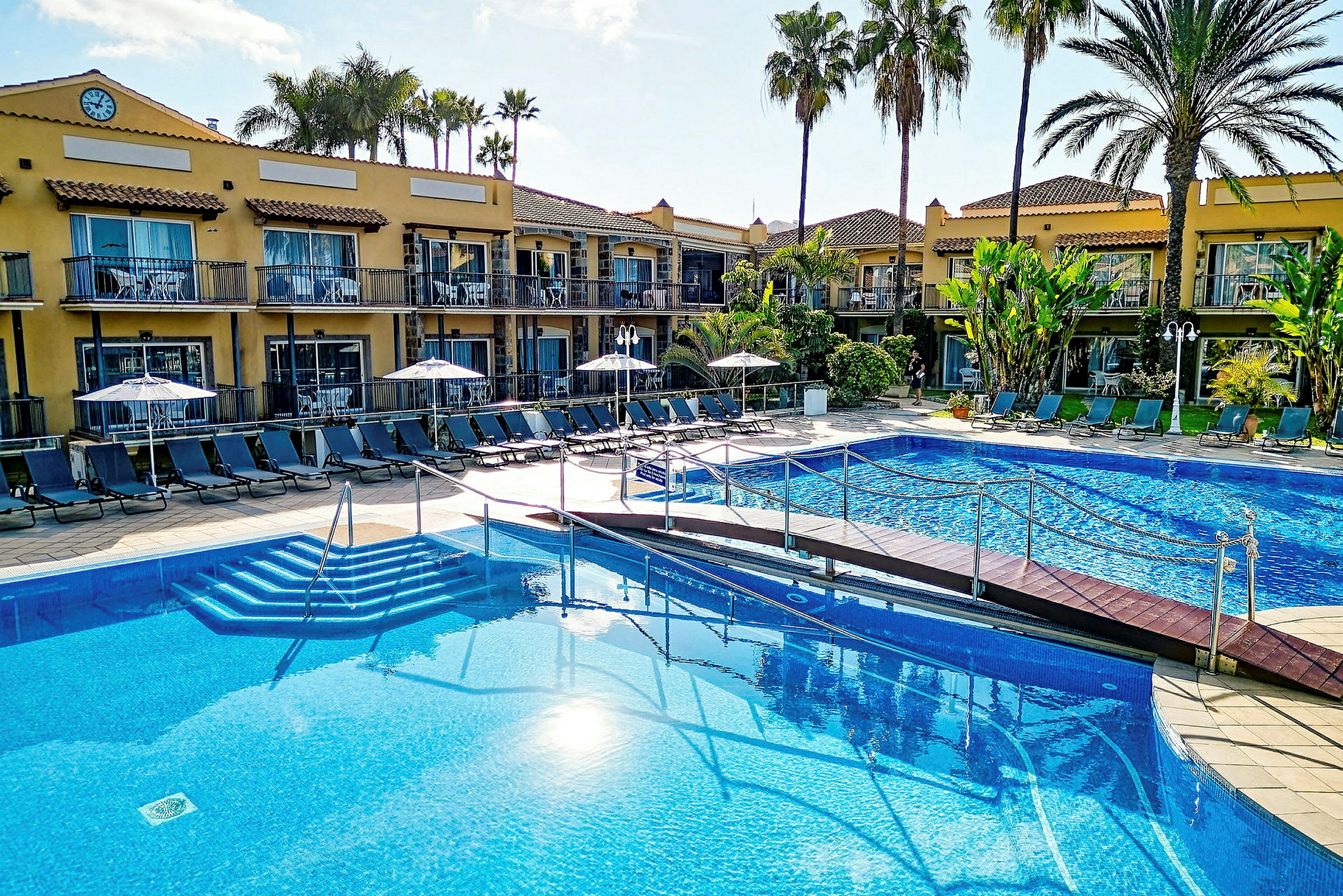 Bild av ett hotell med en pool och solstolar omgivna av palmer. (AI-genererad text)