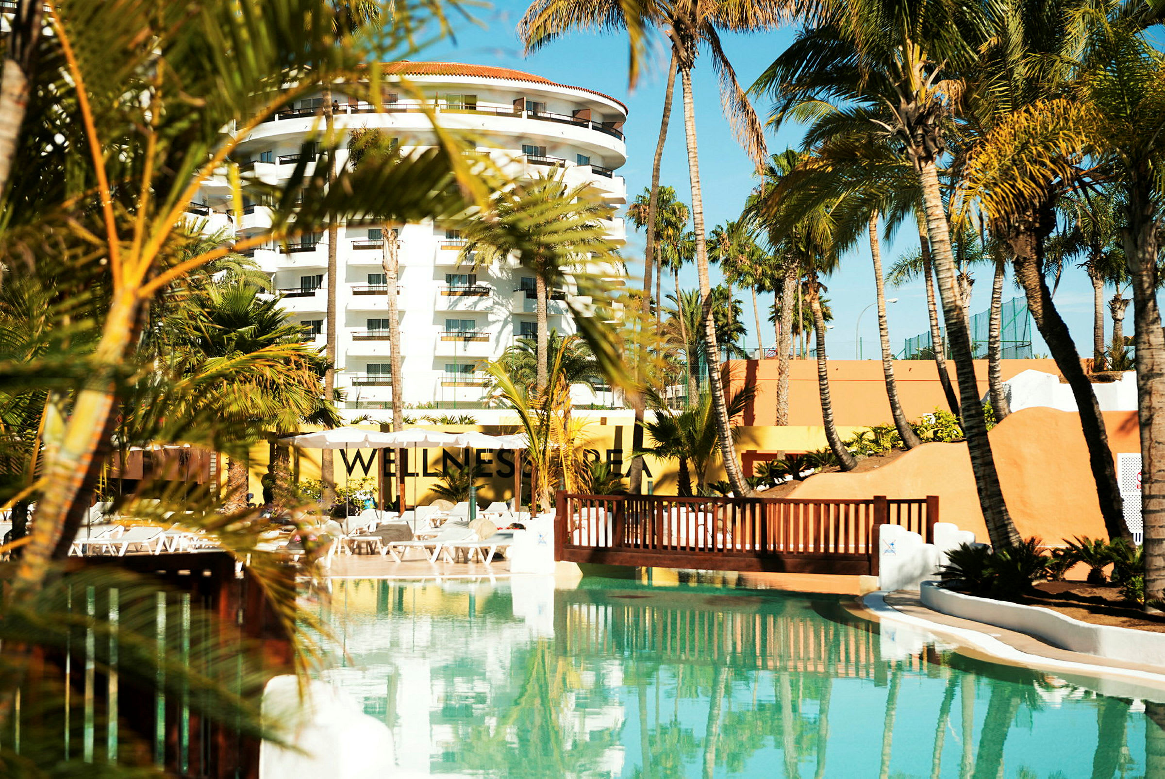 Exteriör av ett hotell med palmträd, pool och texten 'Wellness' synlig. (AI-genererad text)
