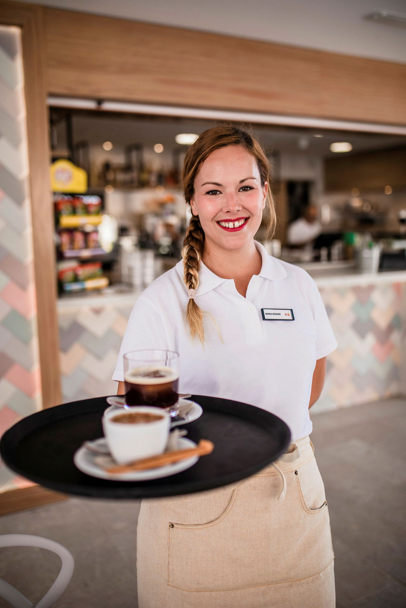 En servitris i uniform håller en bricka med kaffe och te framför en bakgrund av en kafédisk. (AI-genererad text)