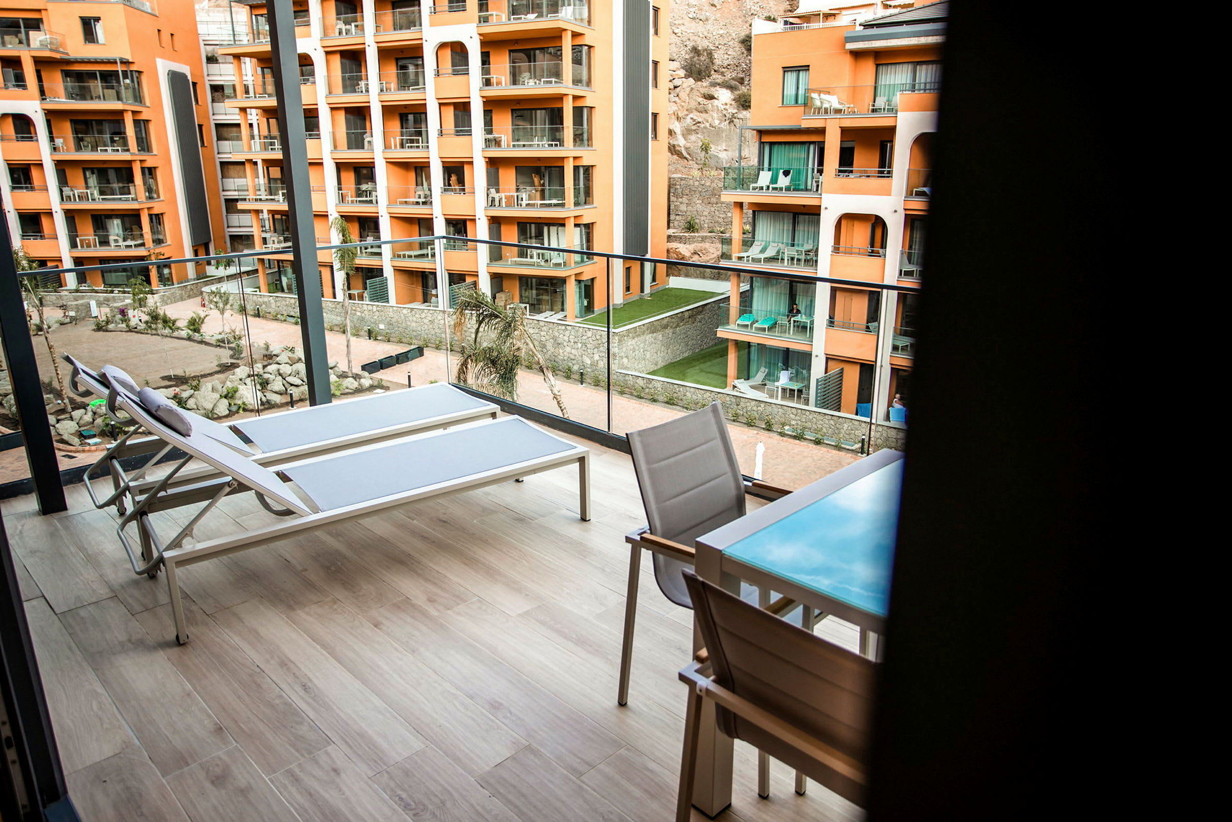 Två solstolar och ett bord med stolar på en rymlig balkong med utsikt över en innergård samt flera orange byggnader. (AI-genererad text)