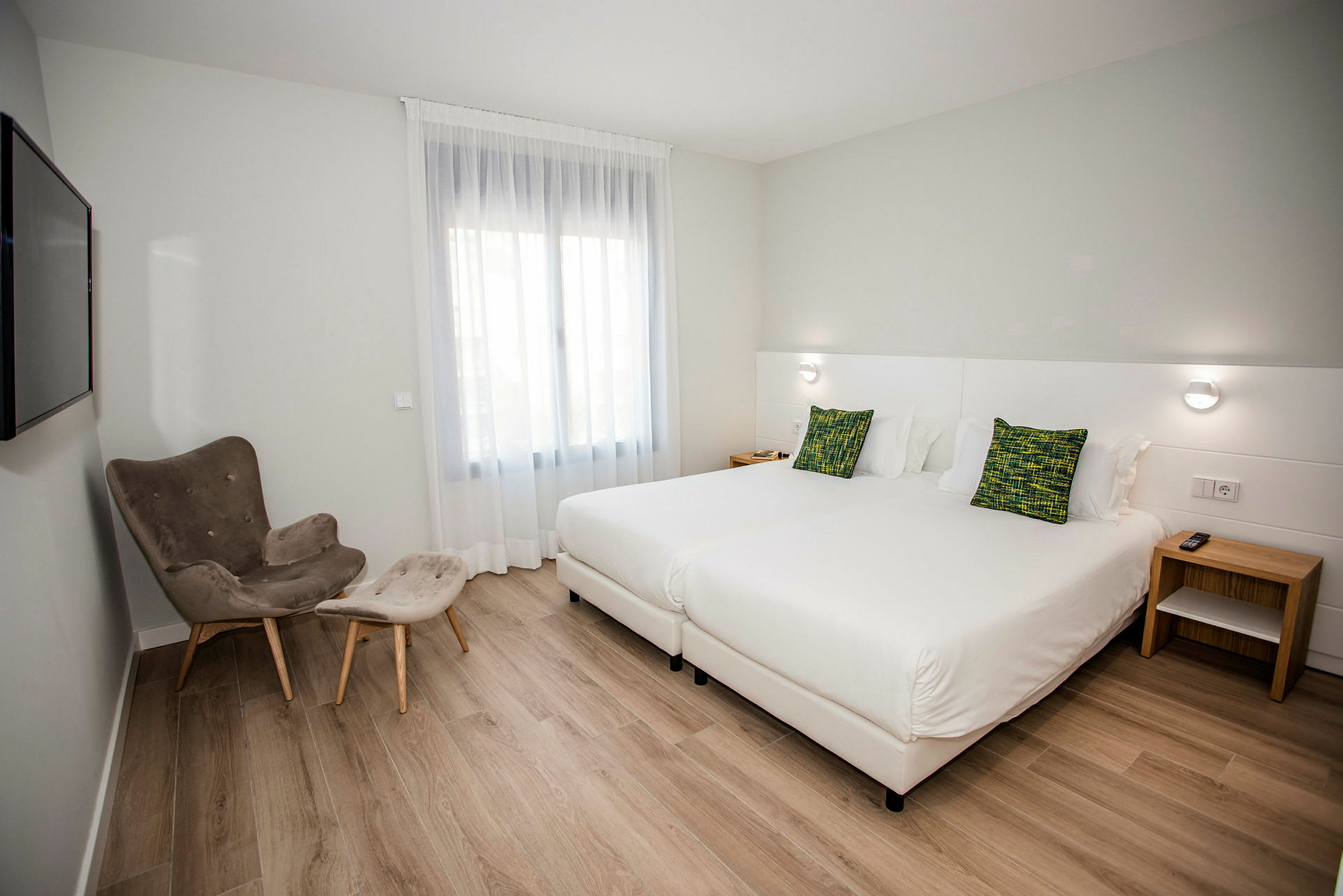 Hotellrum med två enkelsängar, en fåtölj med fotpall samt ett fönster med ljust tyg. (AI-genererad text)