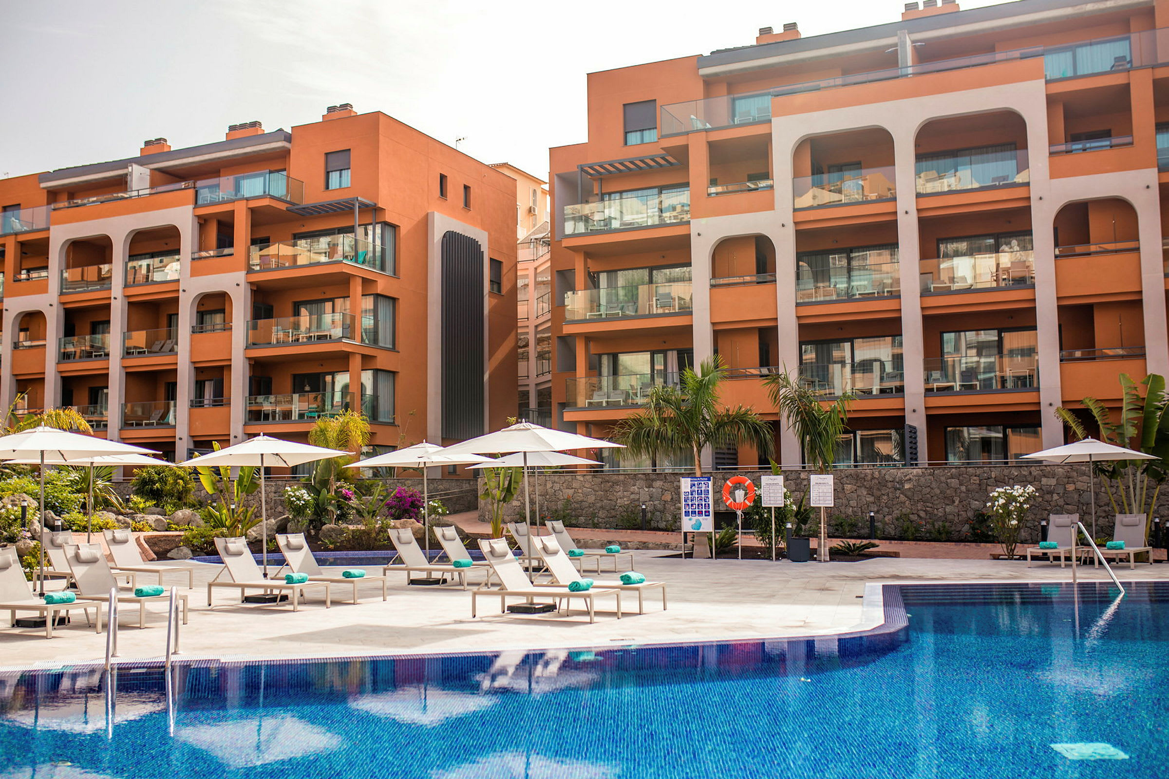 Hotellbyggnad med olika balkonger och en pool framför med solstolar och parasoller utspridda. (AI-genererad text)