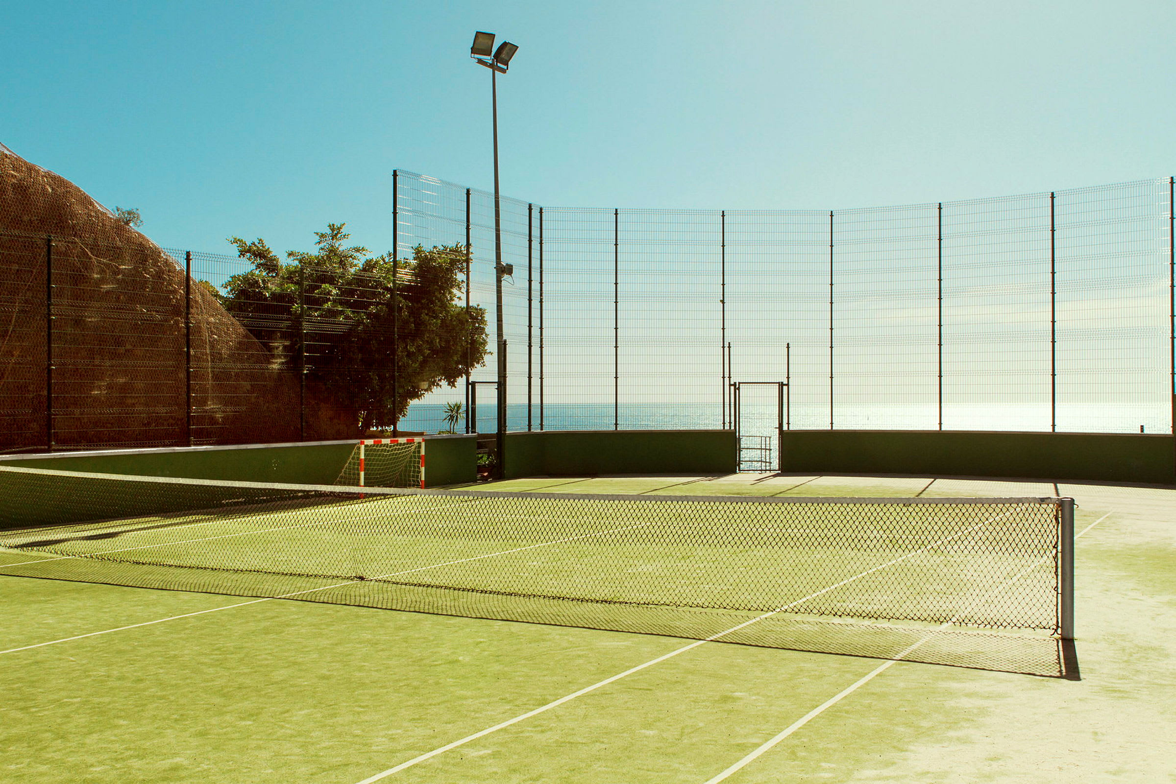 En tennisbana omgiven av nätstängsel nära havet. (AI-genererad text)