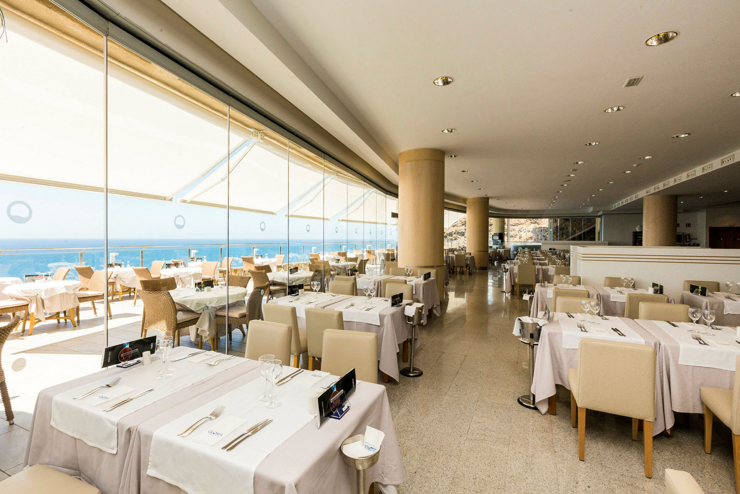En restaurang med bord dukade med vit duk och glas, med utsikt över havet genom stora fönster. (AI-genererad text)