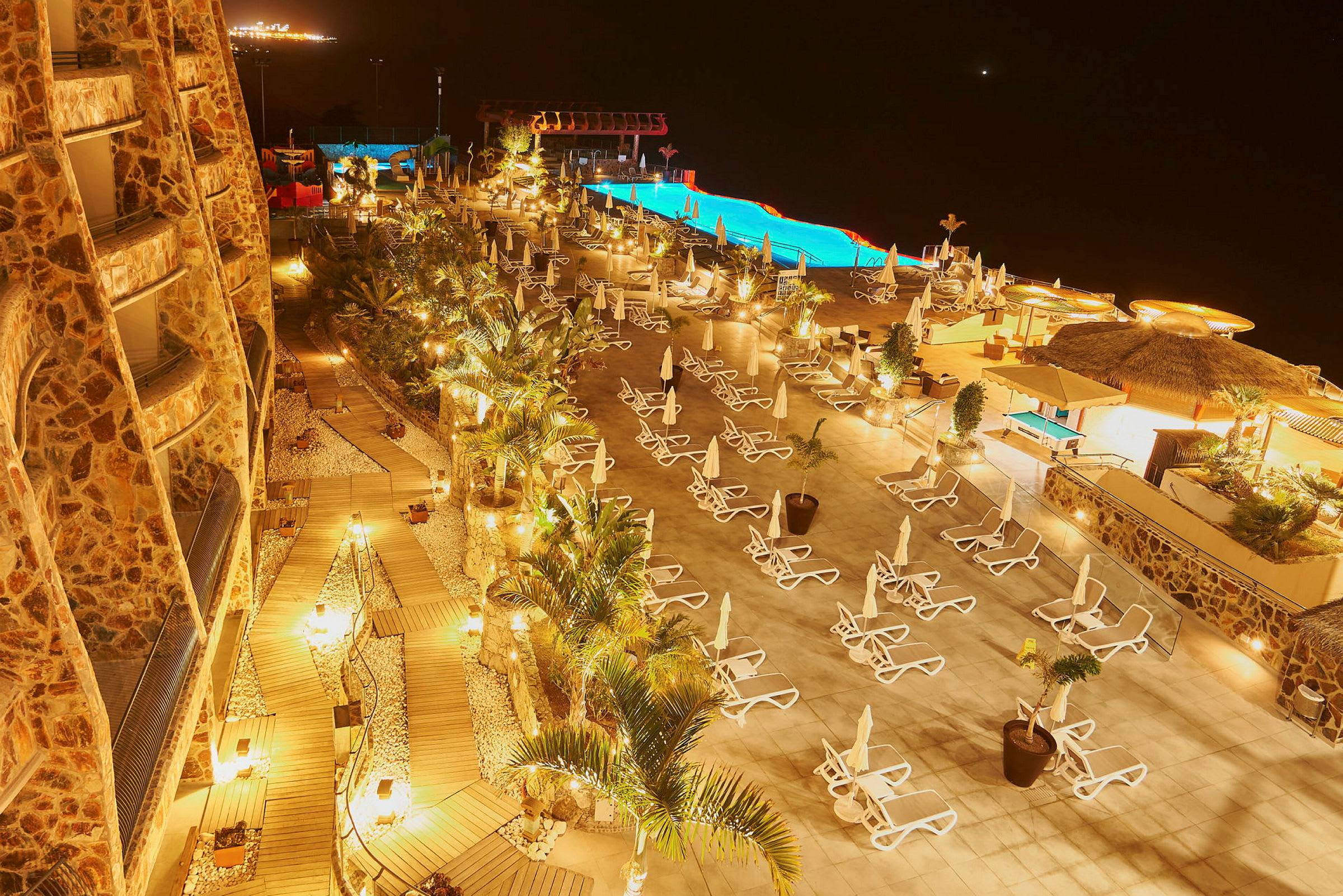 Hotellområde med pool, solstolar och träd, upplyst på natten. (AI-genererad text)