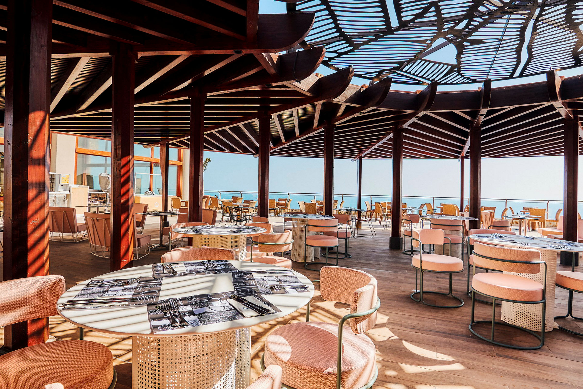 En utomhusrestaurang med solskyddande tak, bord och stolar. Utsikt mot havet. (AI-genererad text)