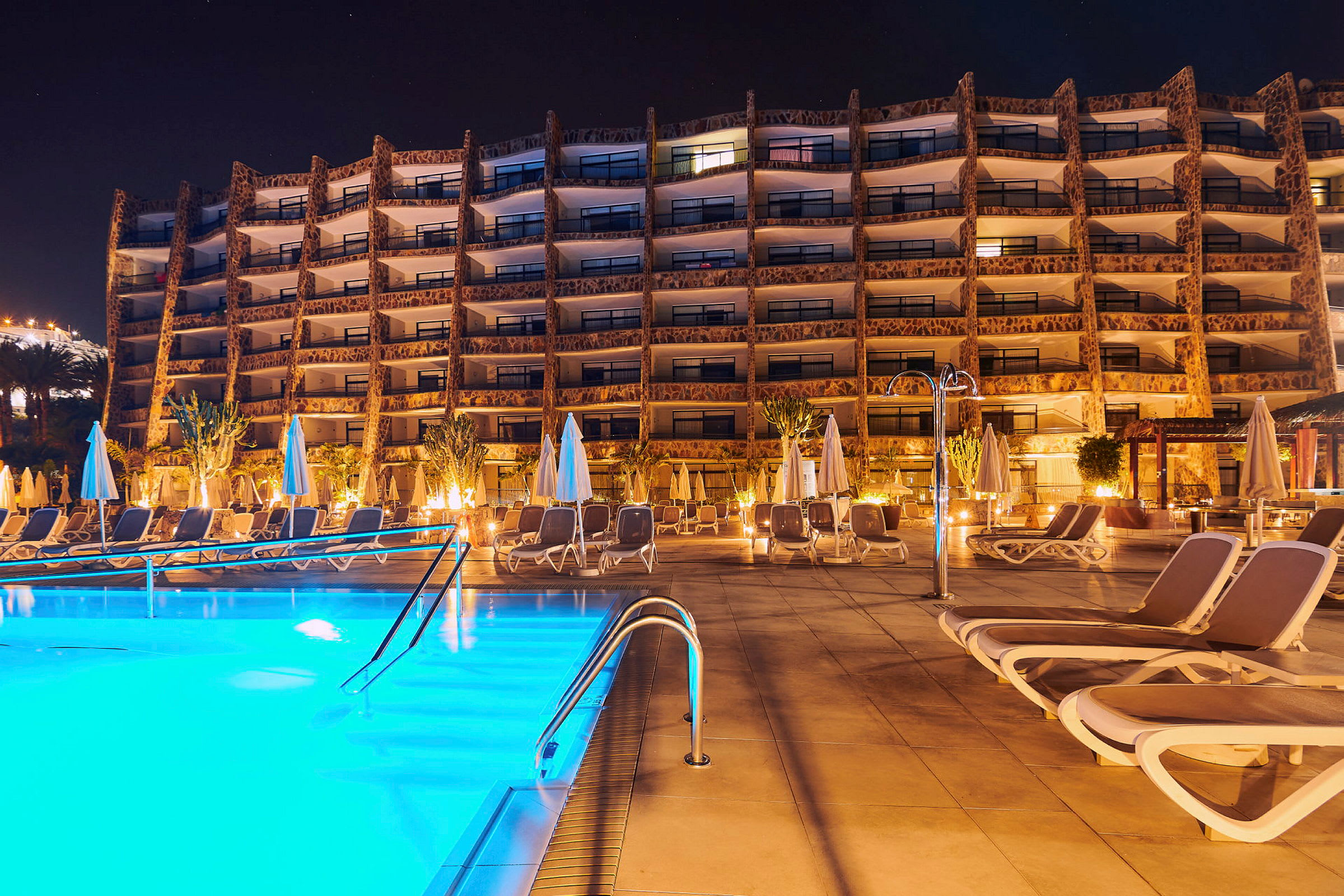 Bild på hotellbyggnad med pool och solstolar i förgrunden, fotograferad på kvällen. (AI-genererad text)
