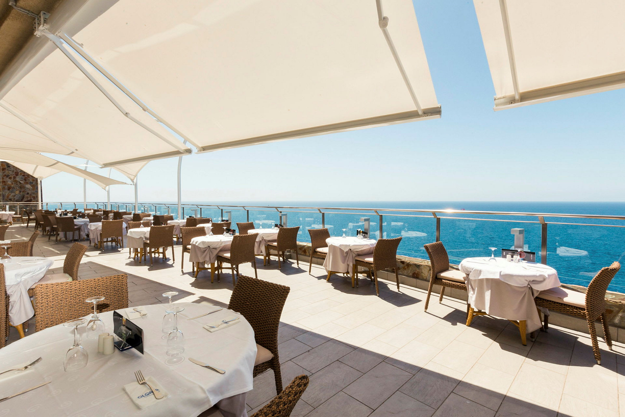 En utomhusrestaurang med havsutsikt och solskydd över sittplatserna. (AI-genererad text)