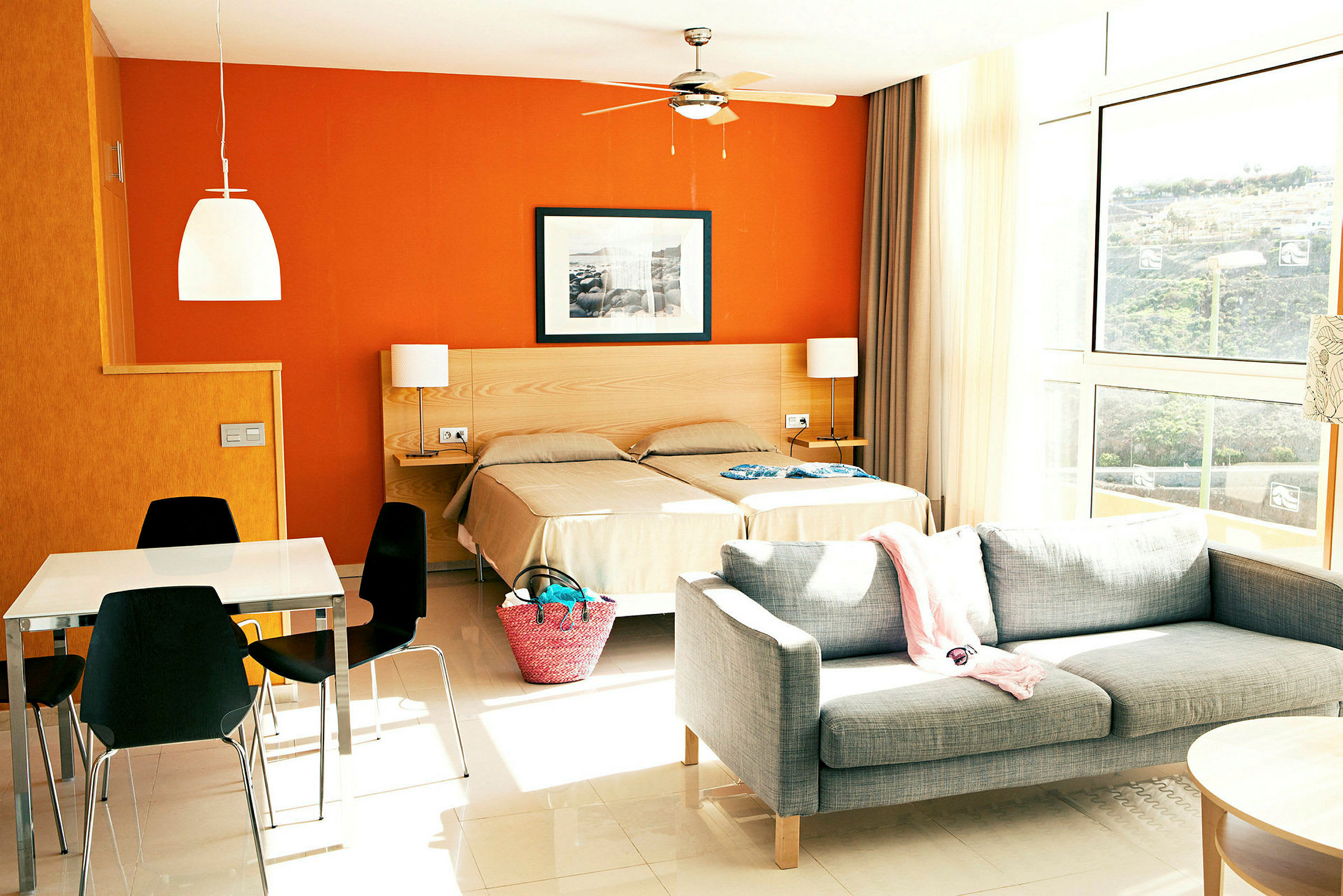 Bild av ett hotellrum med två enkelsängar, grå soffa och ett matbord med svarta stolar. Väggarna är orangea och stora fönster släpper in ljus. (AI-genererad text)