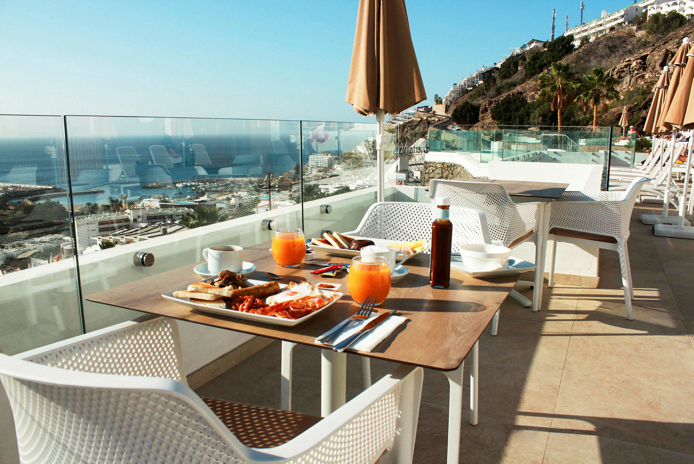 Utsikt över en frukostbord på en terrass med glasräcke, mot havet och en klippa med byggnader. (AI-genererad text)