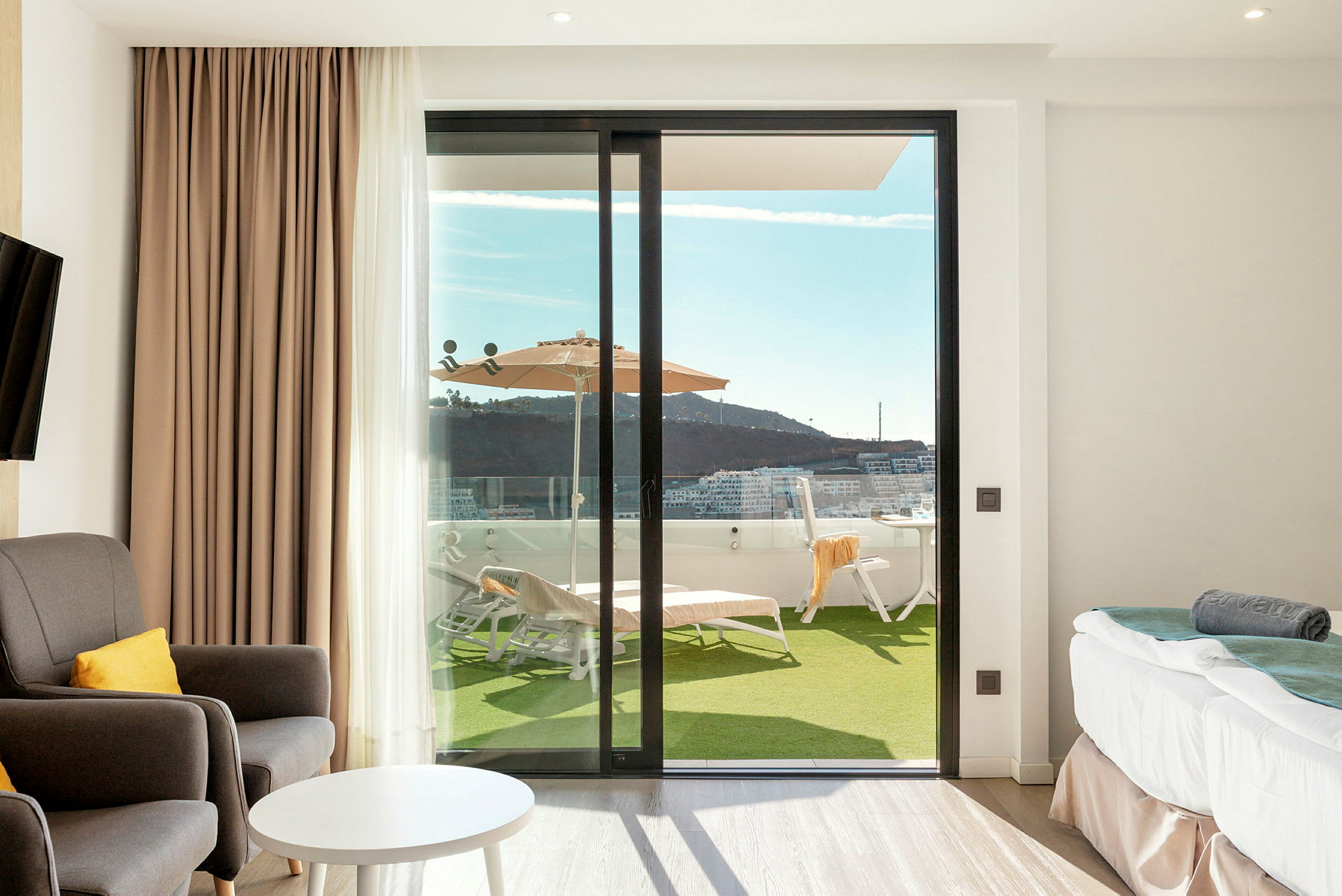 Ett hotellrum med sittmöbler, bord och säng, och en glasdörr som leder ut till en terrass med solstolar och parasoll. (AI-genererad text)