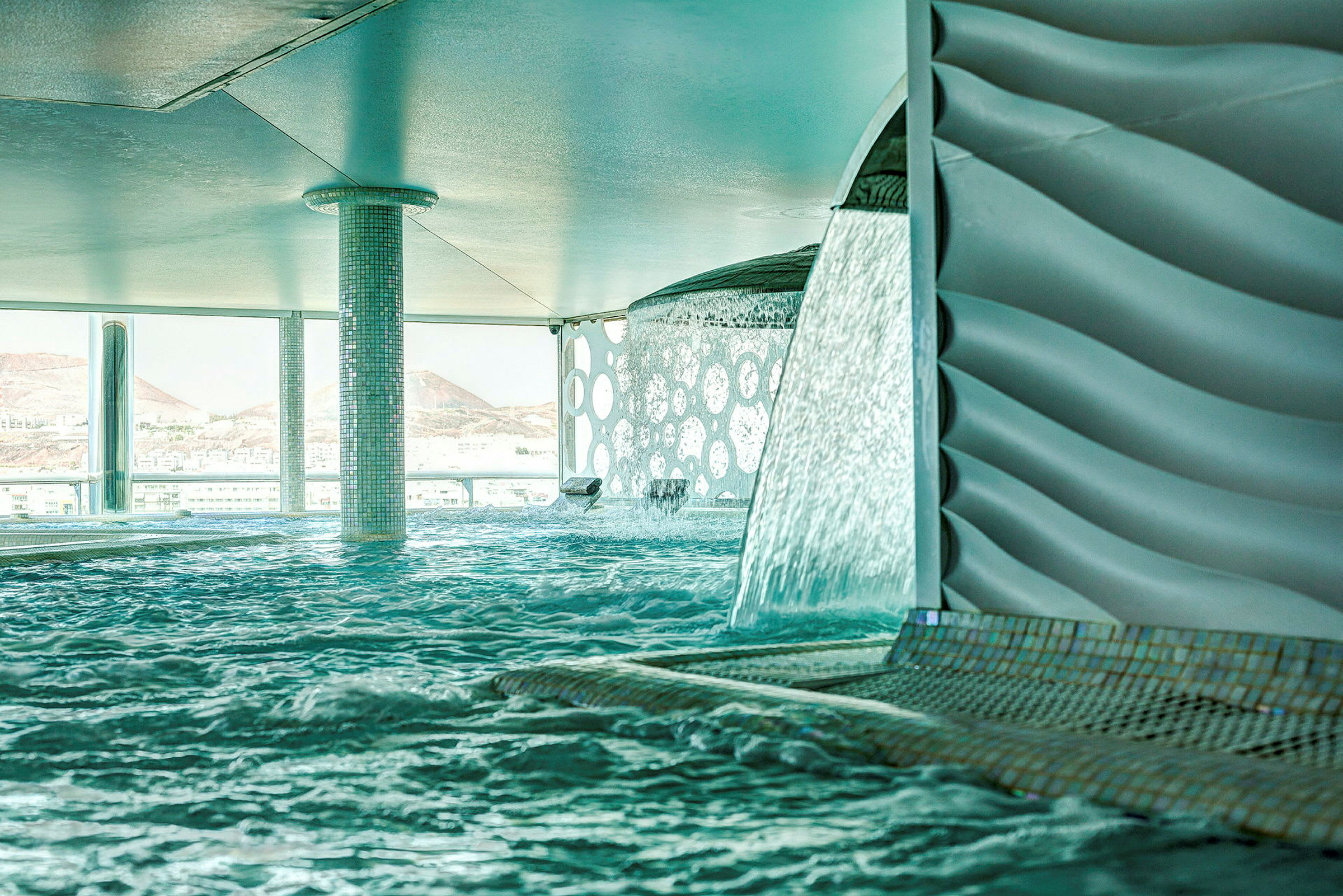 Inomhuspool med vattenfall och utsikt över berg. (AI-genererad text)