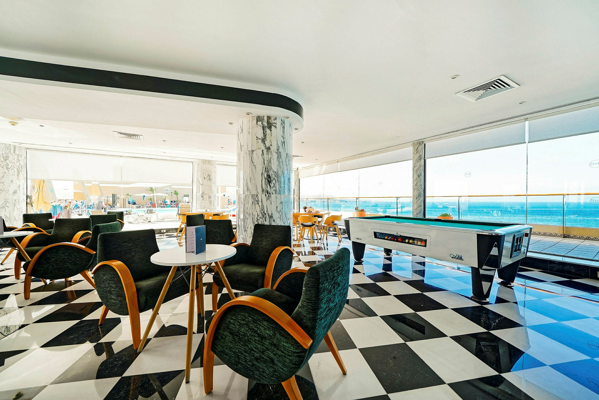 En ljus lounge med sittplatser, biljardbord och utsikt över havet från stora fönster. (AI-genererad text)