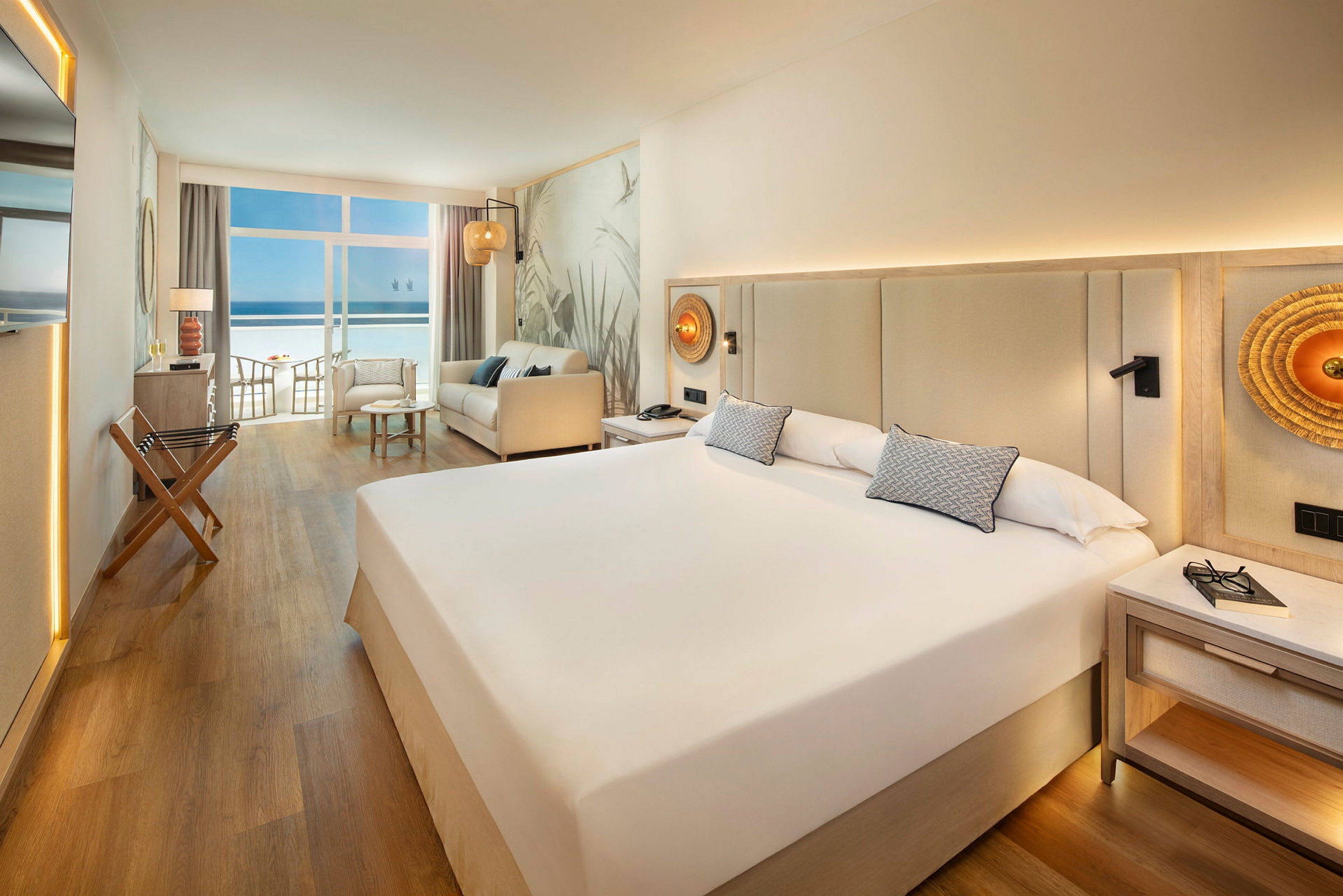 Ett hotellrum med en stor säng, soffa och utsikt över havet genom fönster. (AI-genererad text)