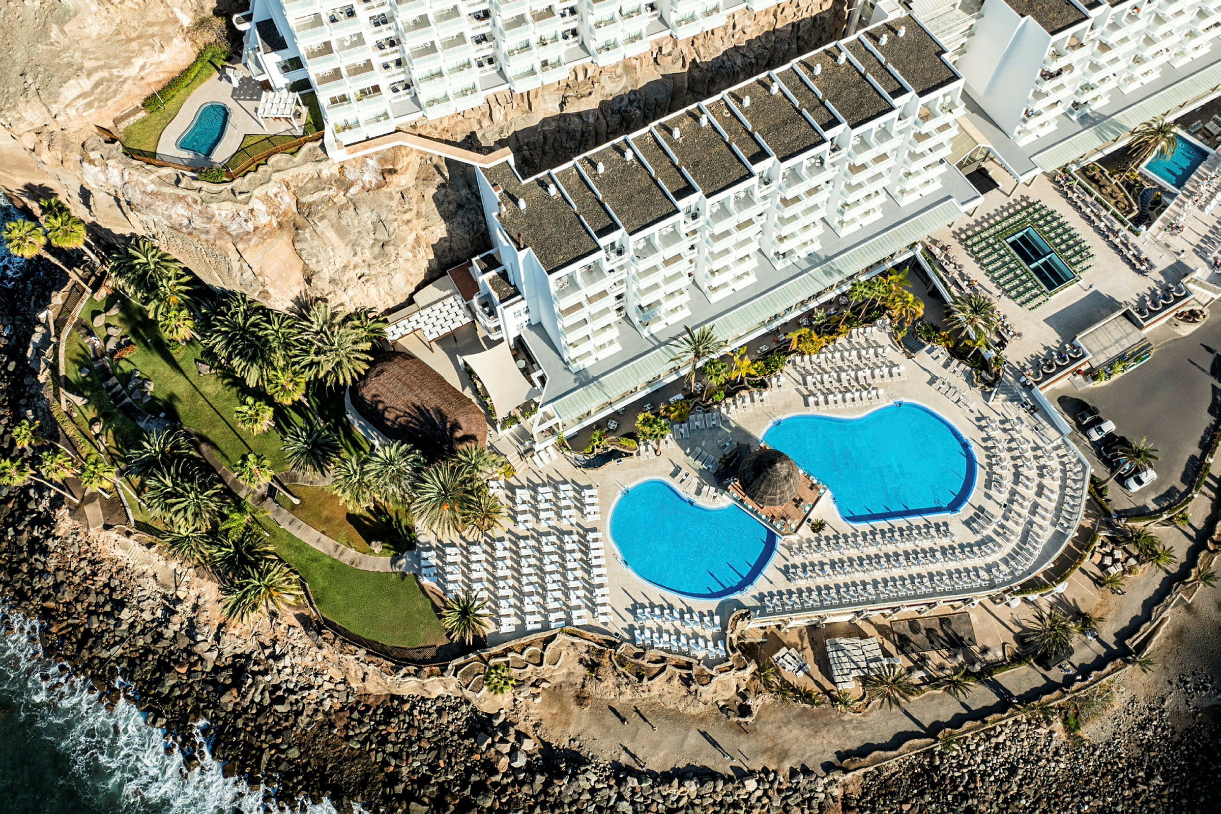 Flygvy över hotellområde med byggnader, pooler, grönområden och kustlinje. (AI-genererad text)
