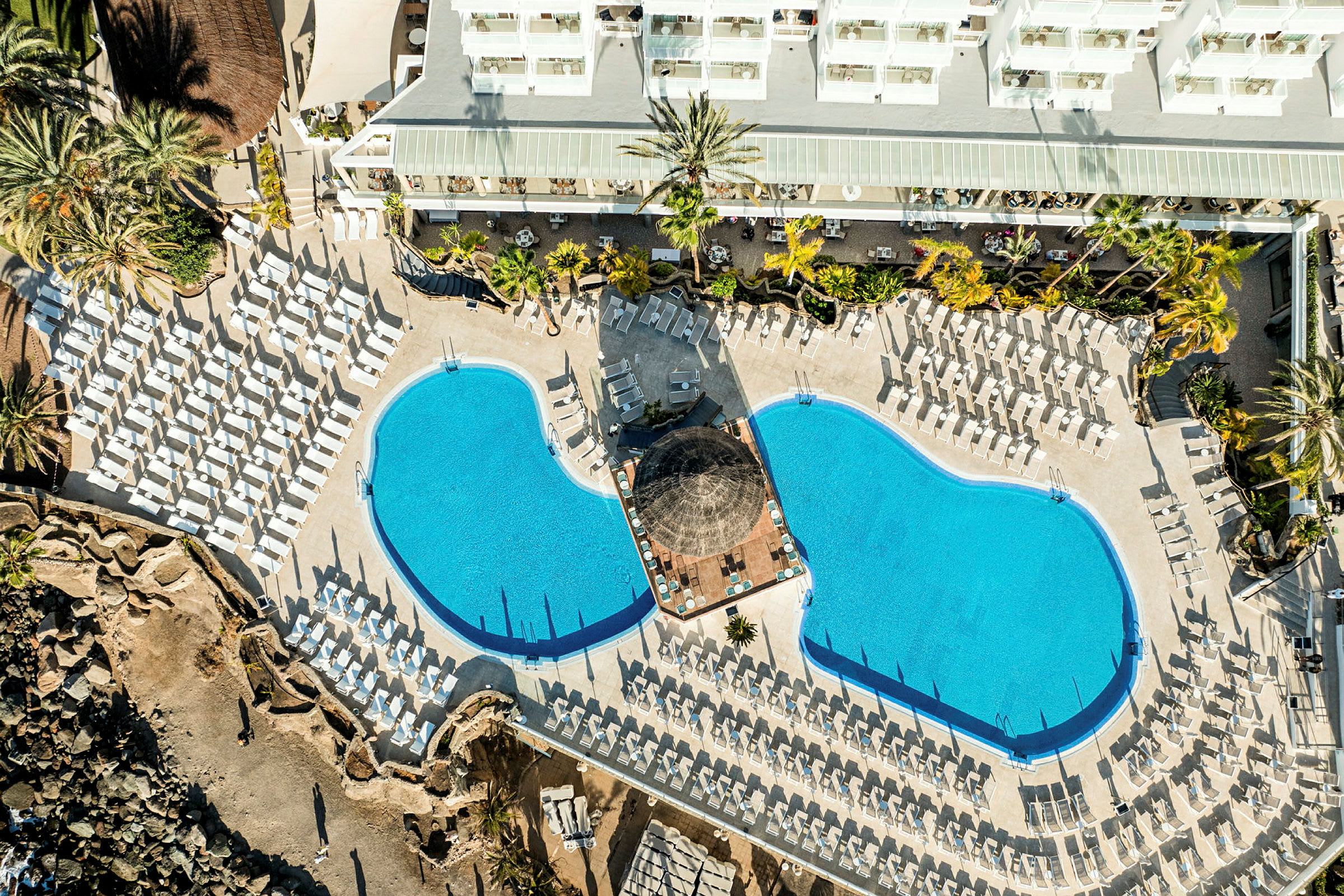 Fågelperspektiv av en hotellets poolområde med två pooler och många solstolar. (AI-genererad text)