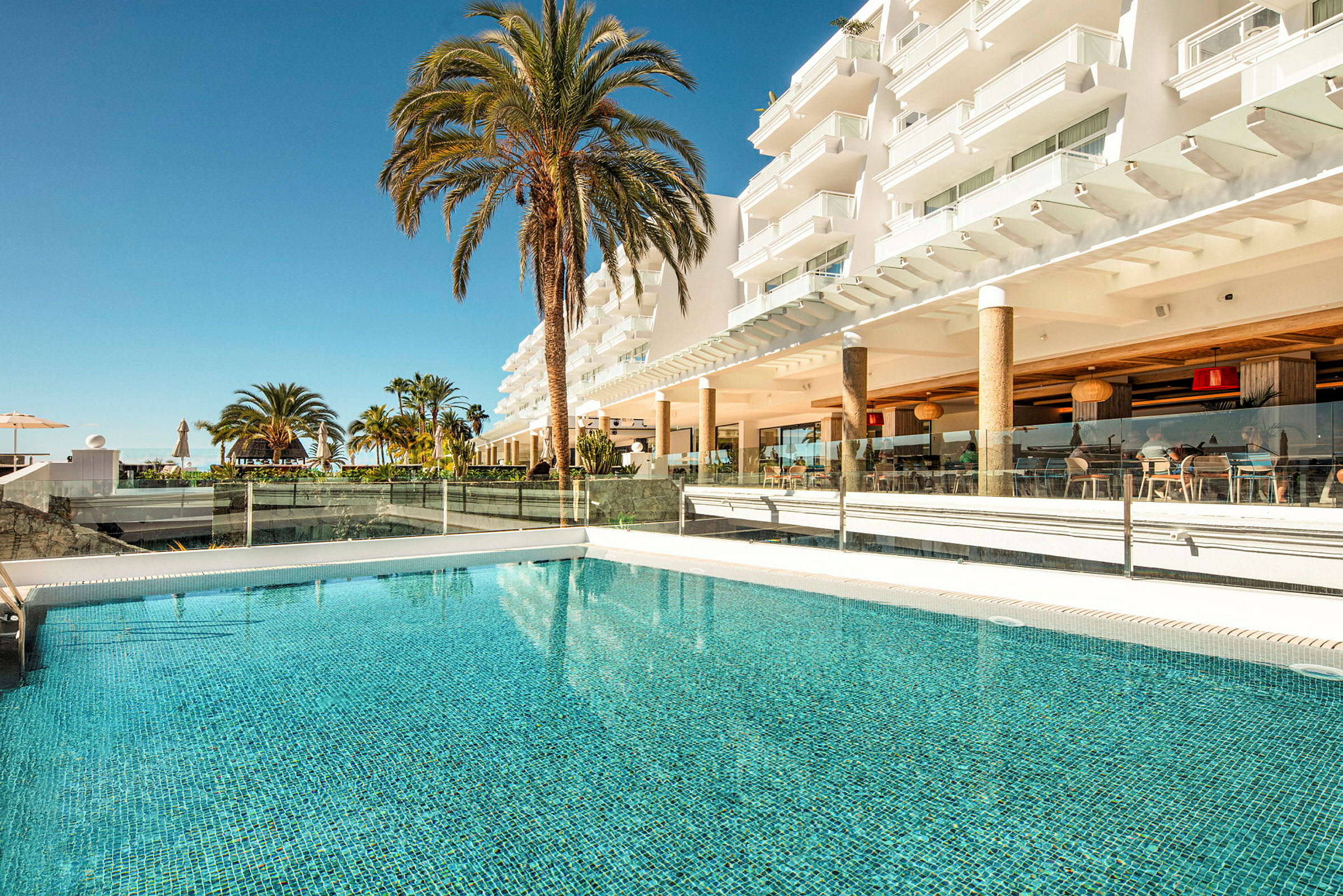 En pool framför ett hotell med balkonger och palmer i bakgrunden. (AI-genererad text)