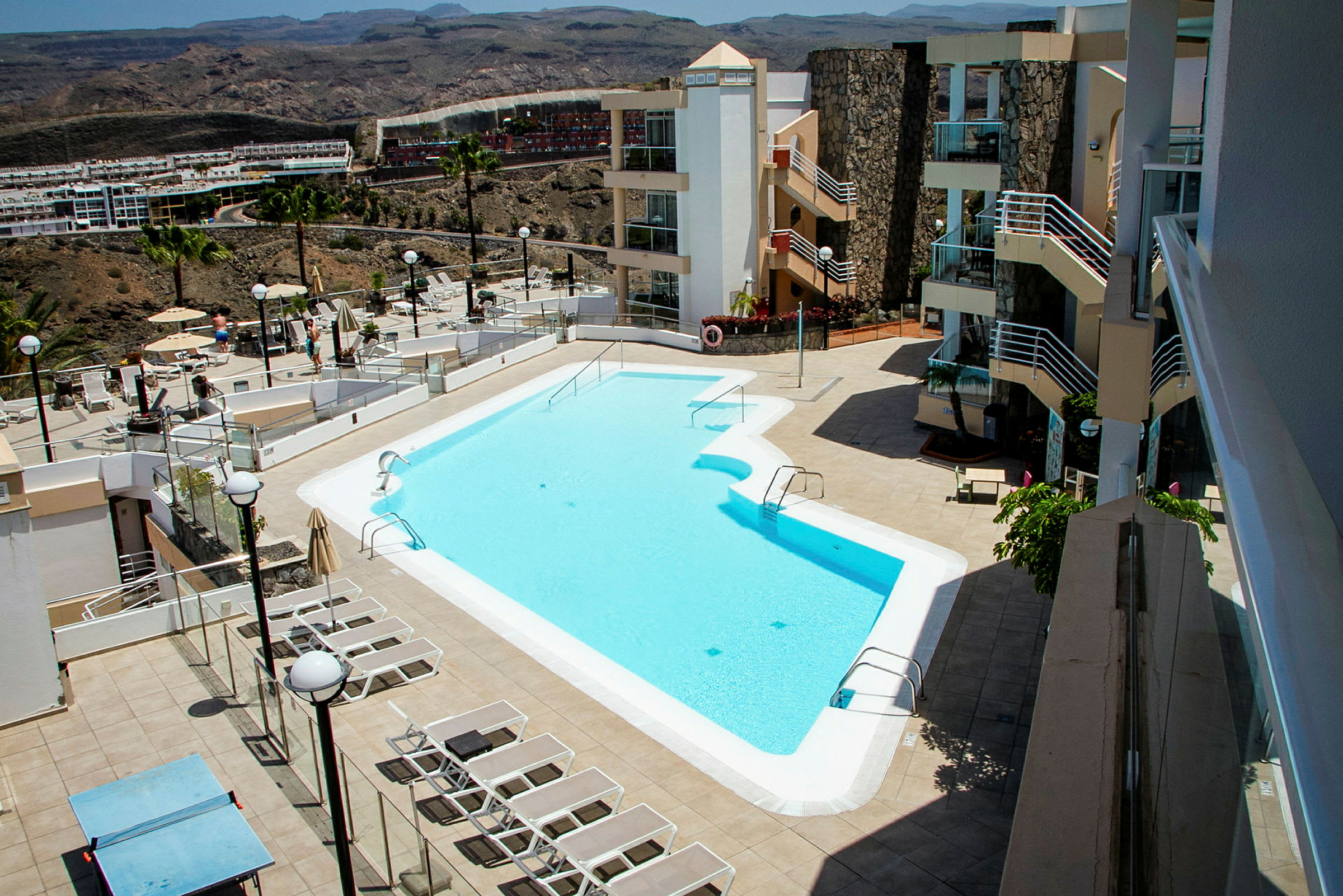 Bild av ett hotellområde med en utomhuspool, solstolar och omgivande byggnader. (AI-genererad text)