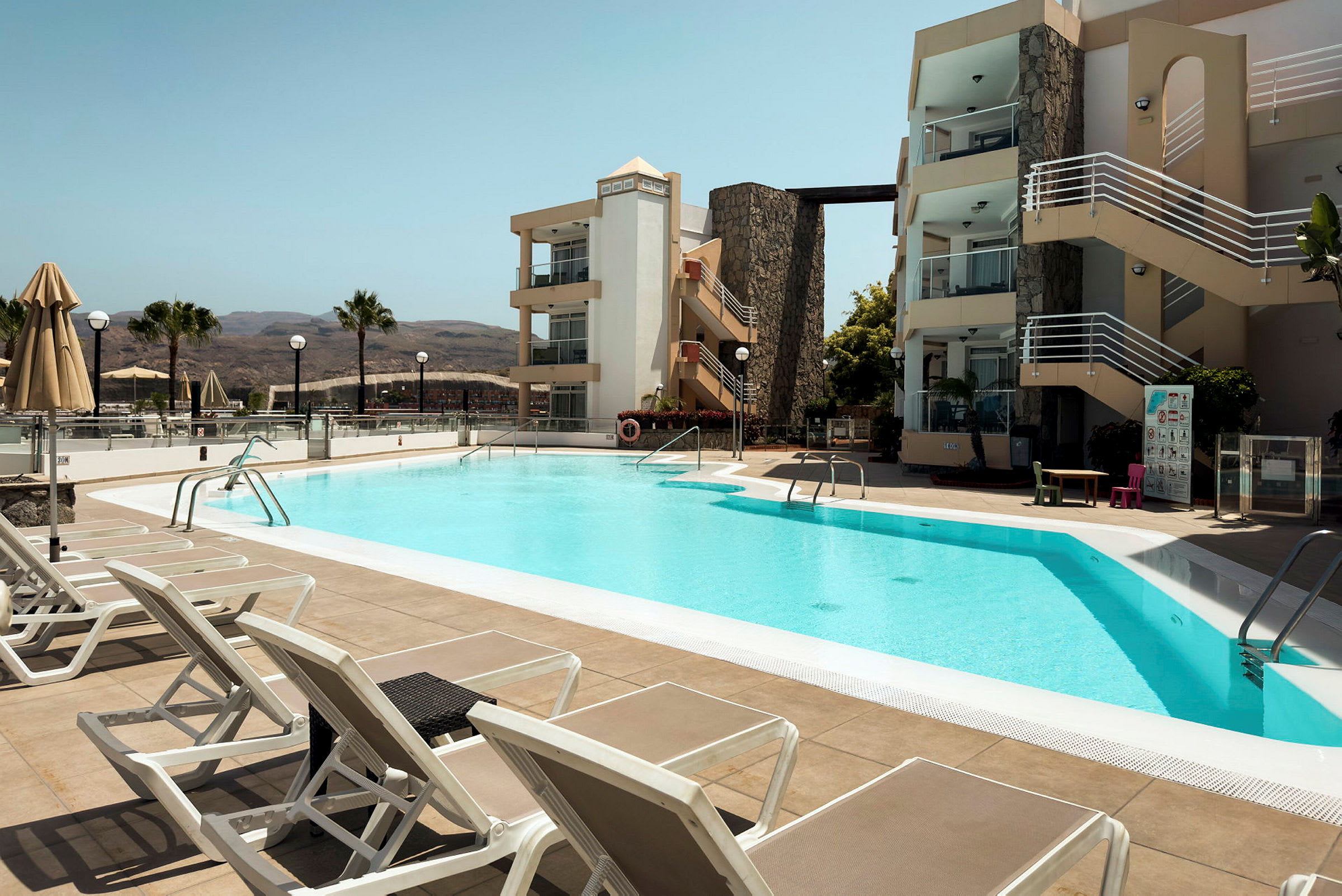 Utomhuspool vid hotell med solstolar och byggnader i bakgrunden. (AI-genererad text)
