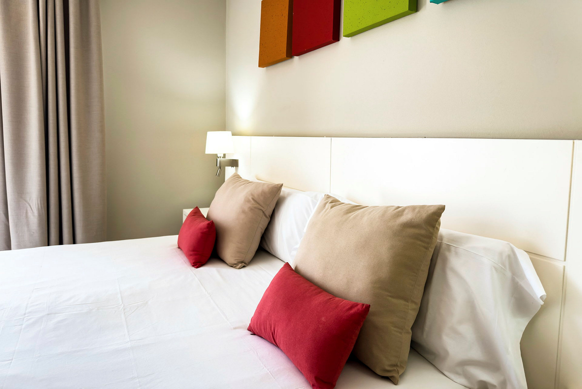 Hotellrum med en säng bäddad med vita lakan och flera kuddar i röda och beige färger. (AI-genererad text)