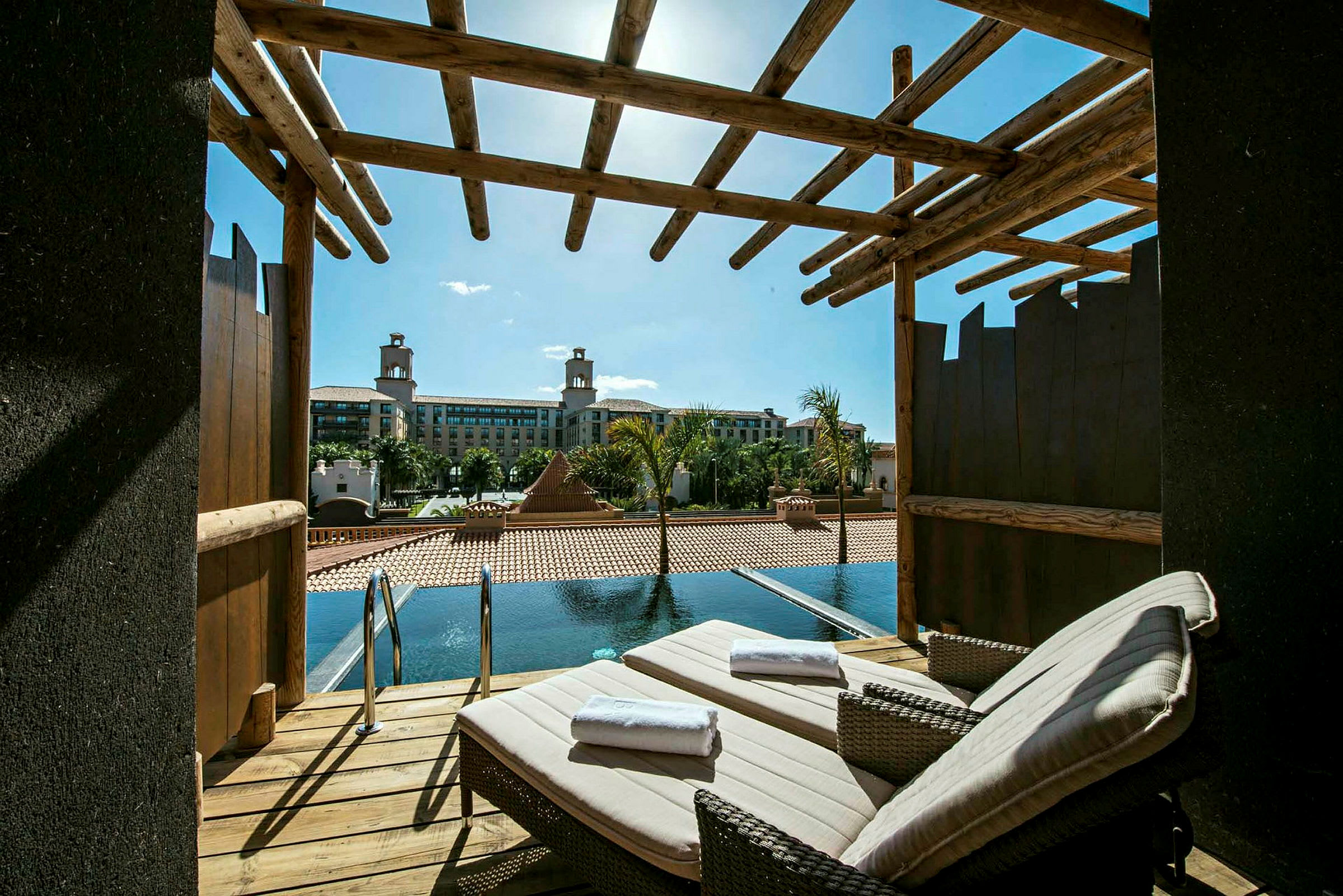 En terrass med två solstolar och utsikt mot en pool och ett hotell i bakgrunden. (AI-genererad text)