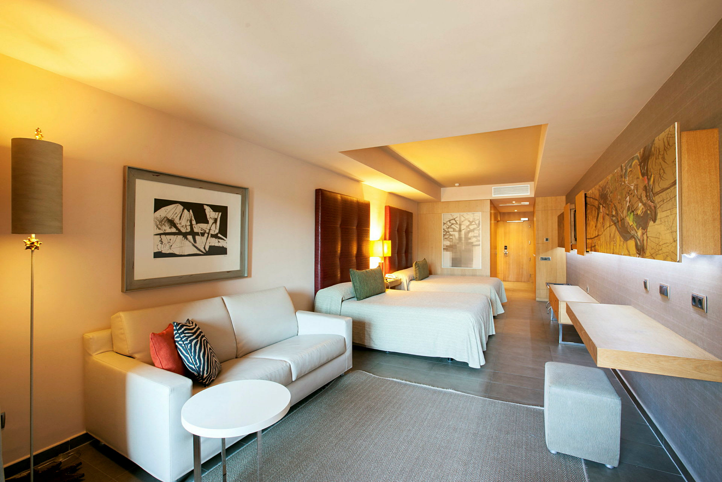 Ett hotellrum med en soffa, två sängar och ett bord, inrett i varm belysning. (AI-genererad text)