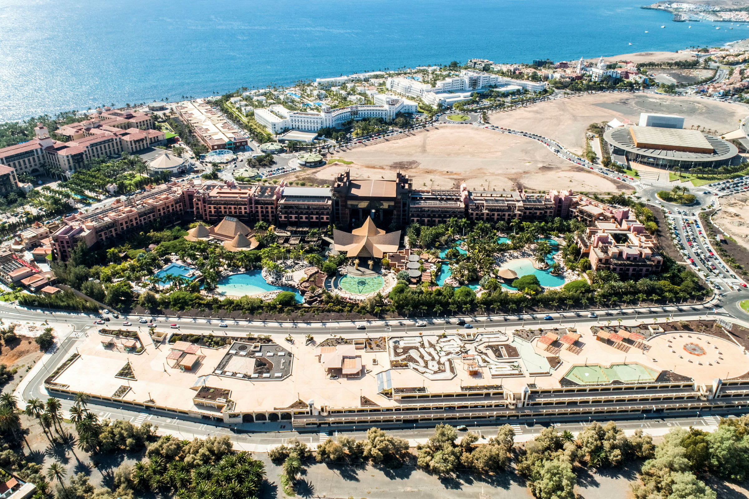 Flygfoto av ett hotellkomplex med pooler, omgivande trädgård och närliggande byggnader vid havet. (AI-genererad text)