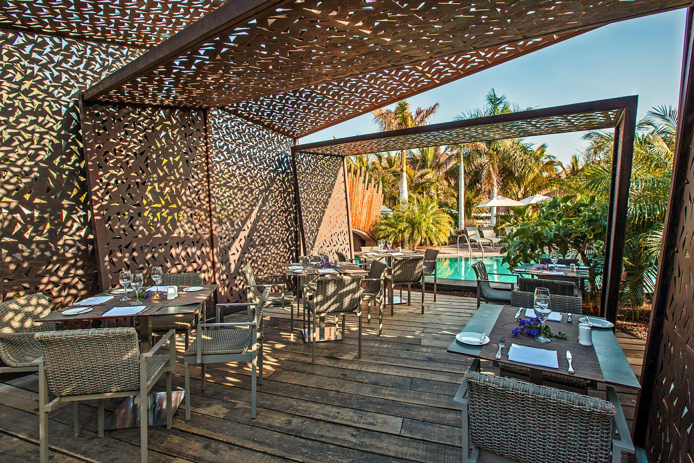 En utomhusrestaurang med bord och stolar under en skuggande struktur, bredvid en pool och omgiven av palmer. (AI-genererad text)
