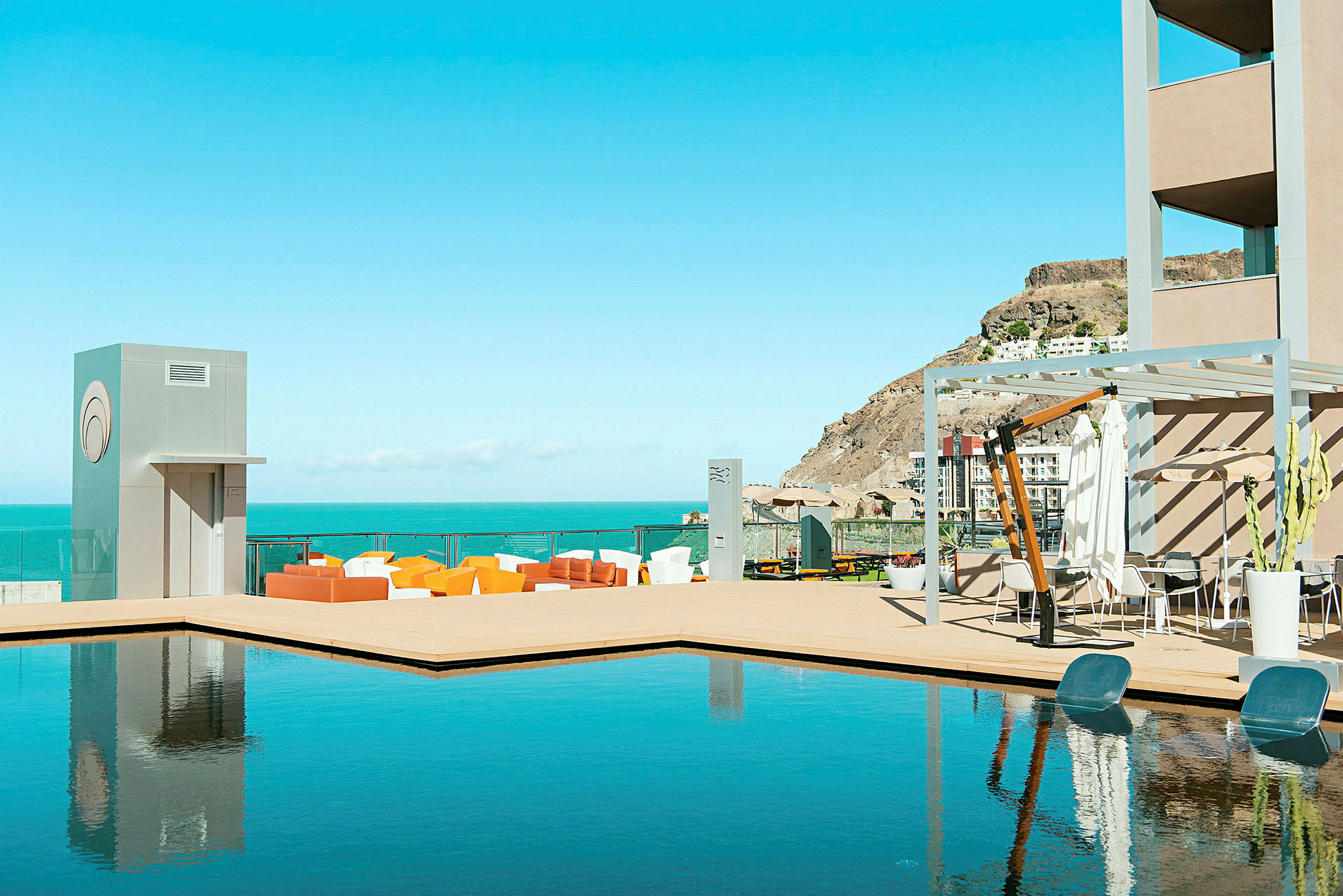 Bild som visar en terrass med pool och sittmöbler. Havet och kullar syns i bakgrunden. (AI-genererad text)