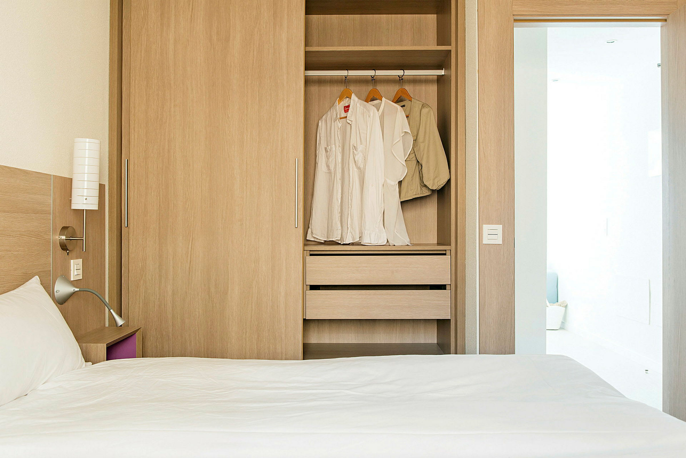 Ett hotellrum med en säng och en garderob som har öppna dörrar och kläder hängande i. (AI-genererad text)