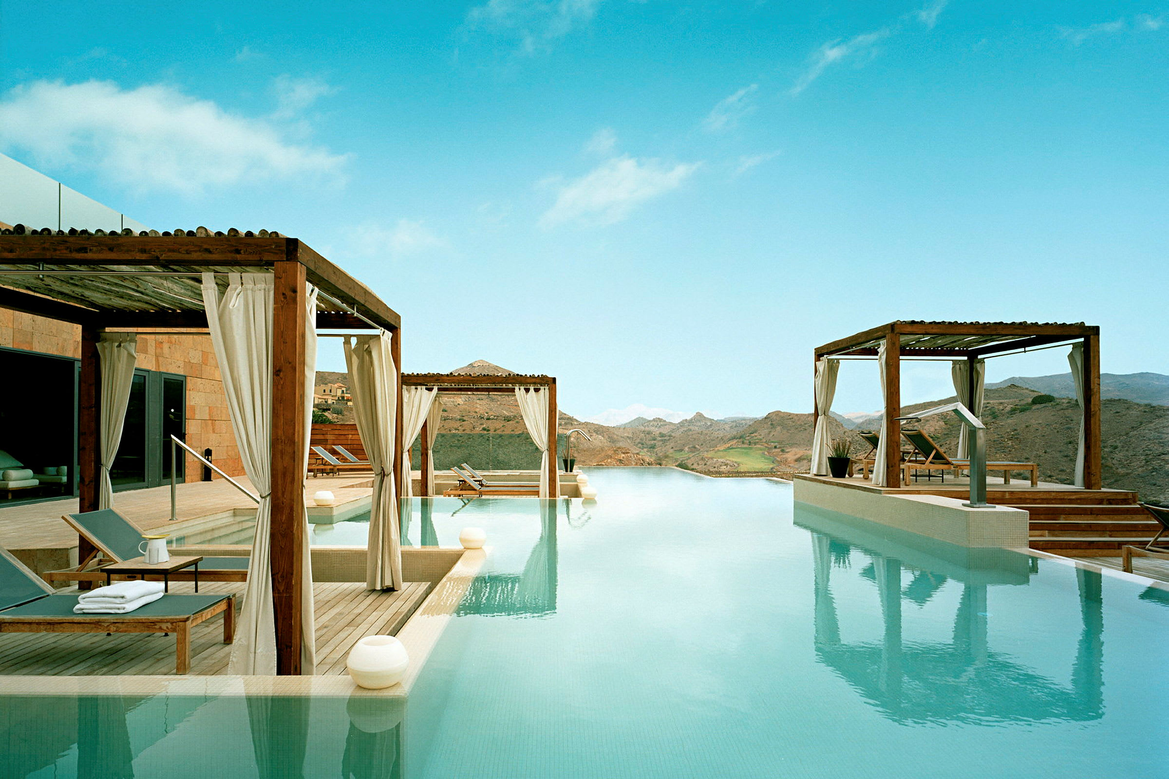 Infinity-pool med utsikt över berglandskapet och omgivande terrasser med solstolar under skärmtak. (AI-genererad text)