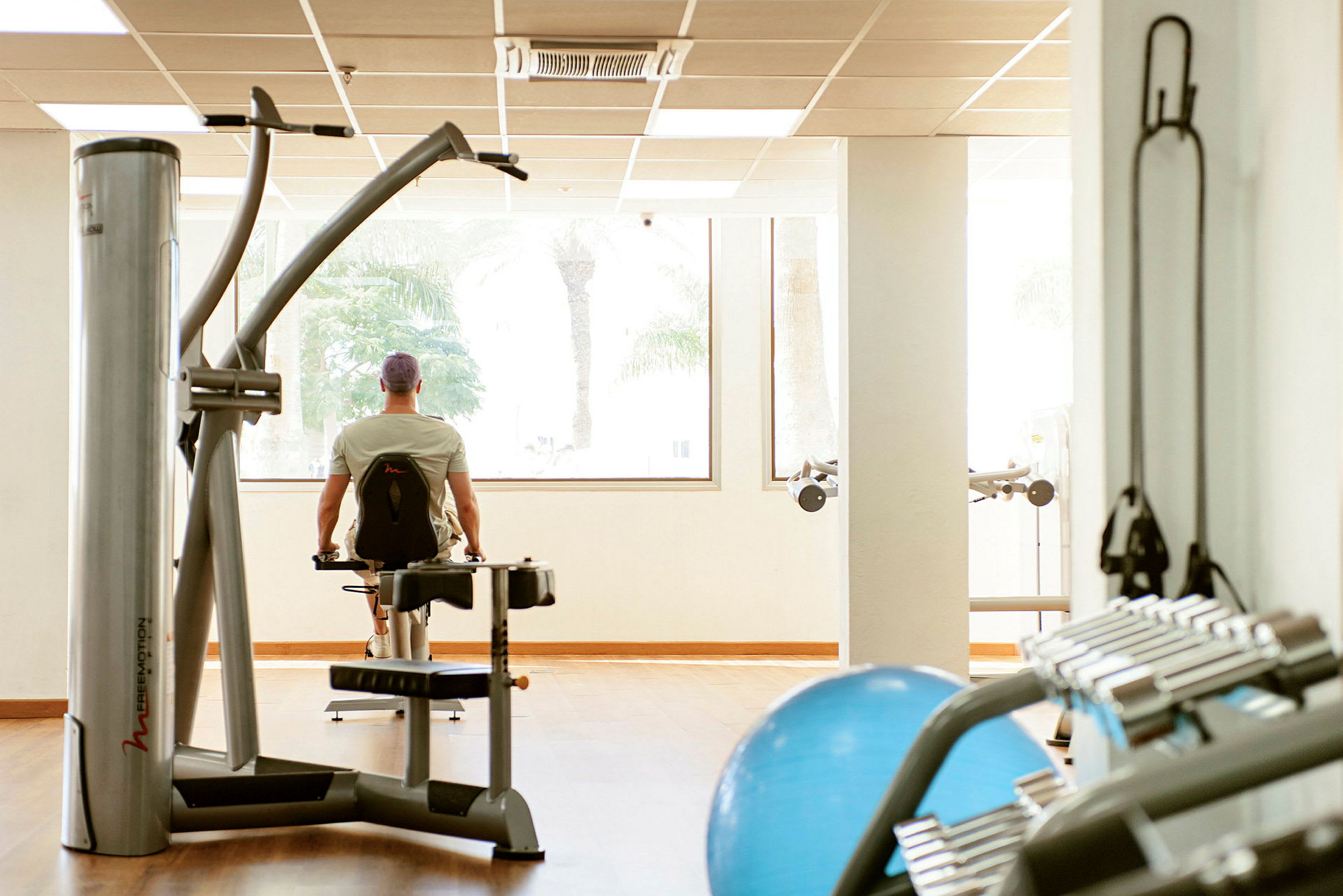 En gymmiljö med träningsutrustning och en person som använder en maskin framför fönster. (AI-genererad text)