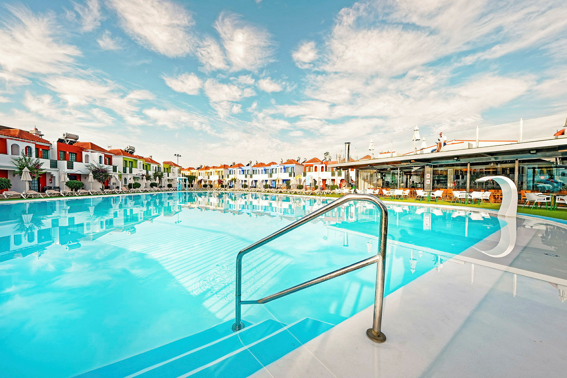 Utomhuspool omgiven av färgglada byggnader och parasoll vid lugnt väder. (AI-genererad text)