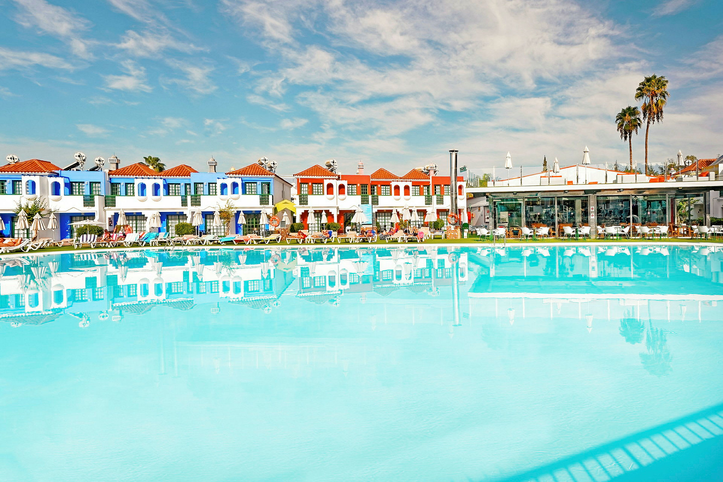 En swimmingpool framför en rad färgglada byggnader och palmer under en klar blå himmel. (AI-genererad text)