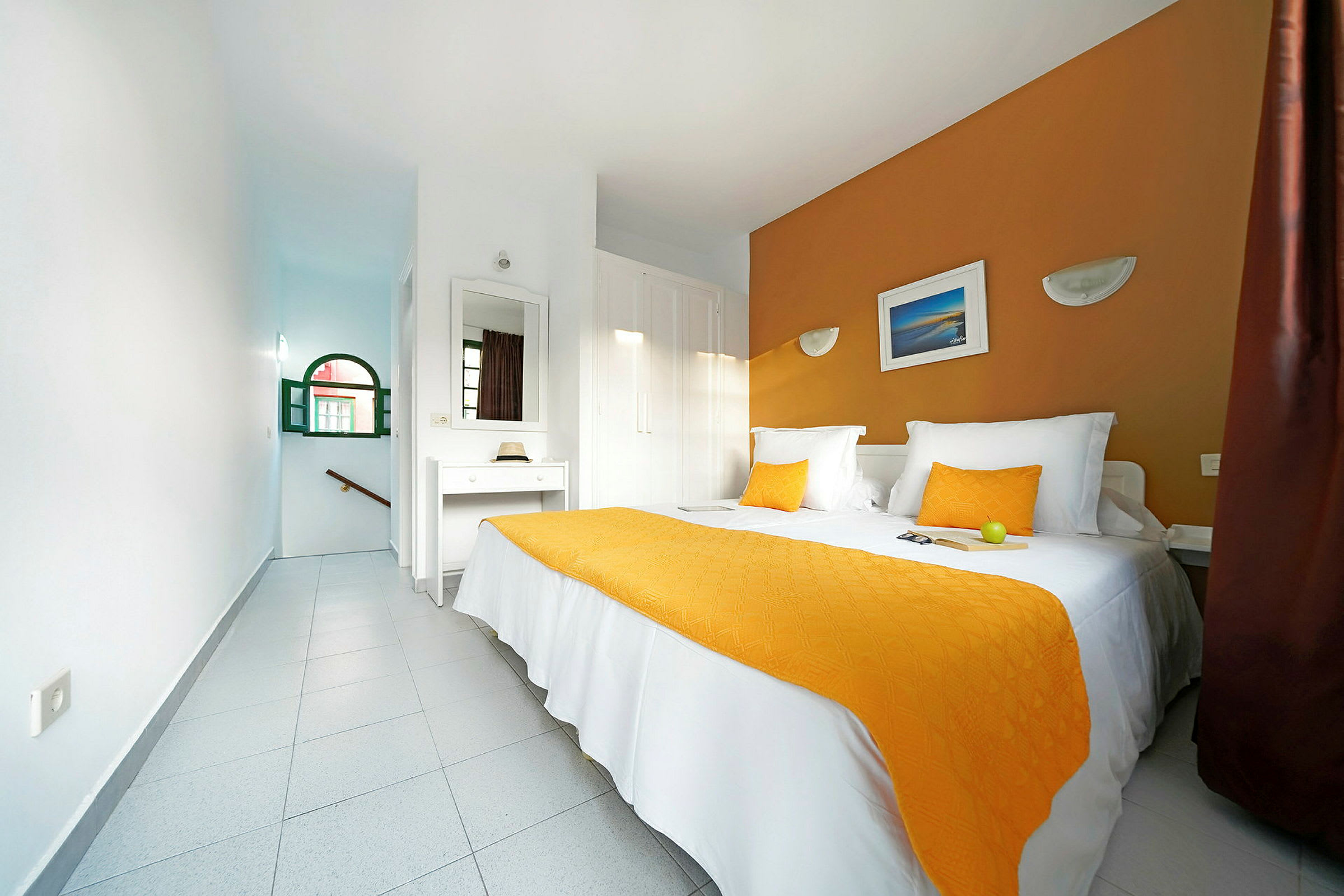 Ett hotellrum med en dubbelsäng klädd med vita sängkläder och orange täcke, spegel på väggen och fönster med grön karm. (AI-genererad text)