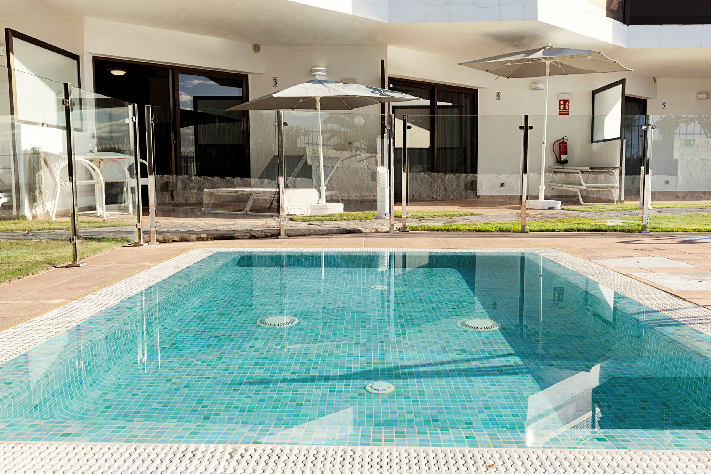 Utomhuspool med glasstaket och omgivande gräsmatta framför en hotellbyggnad. (AI-genererad text)