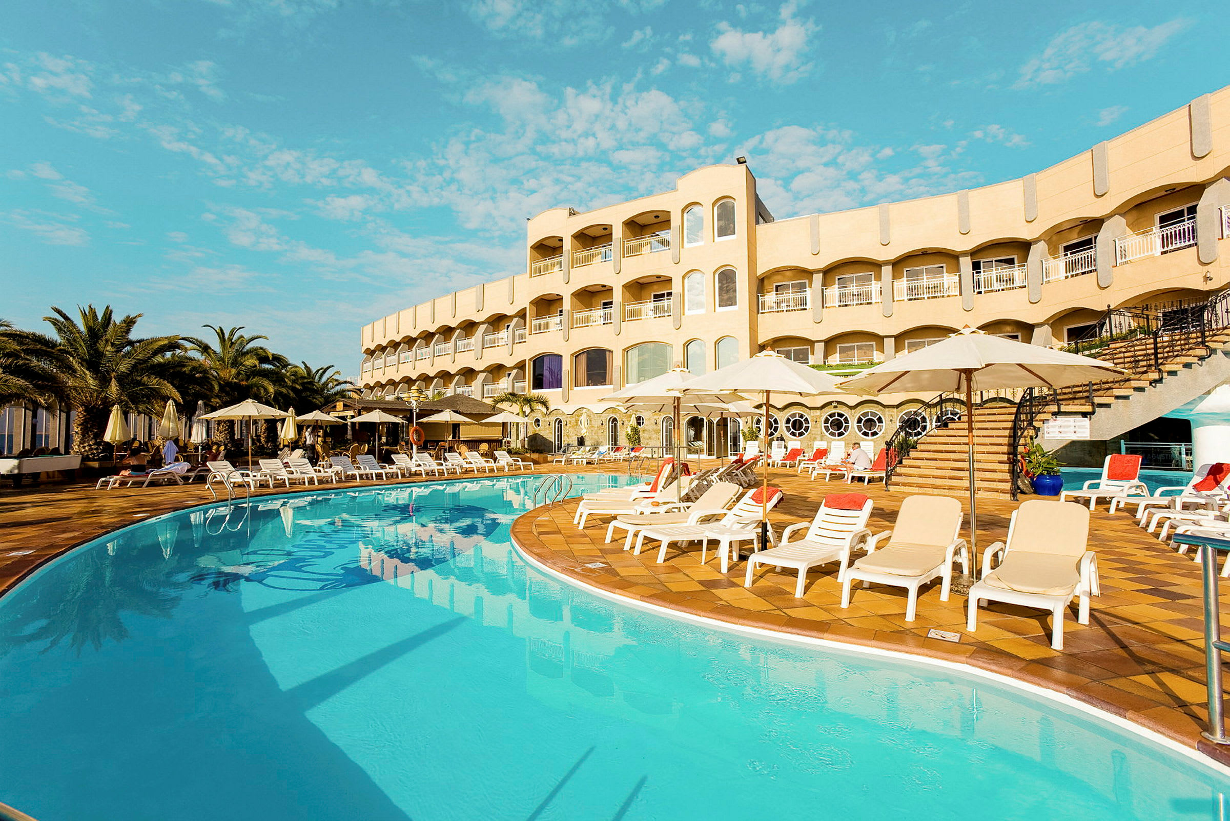 Utomhuspool med solstolar och parasoll framför en hotellbyggnad. (AI-genererad text)