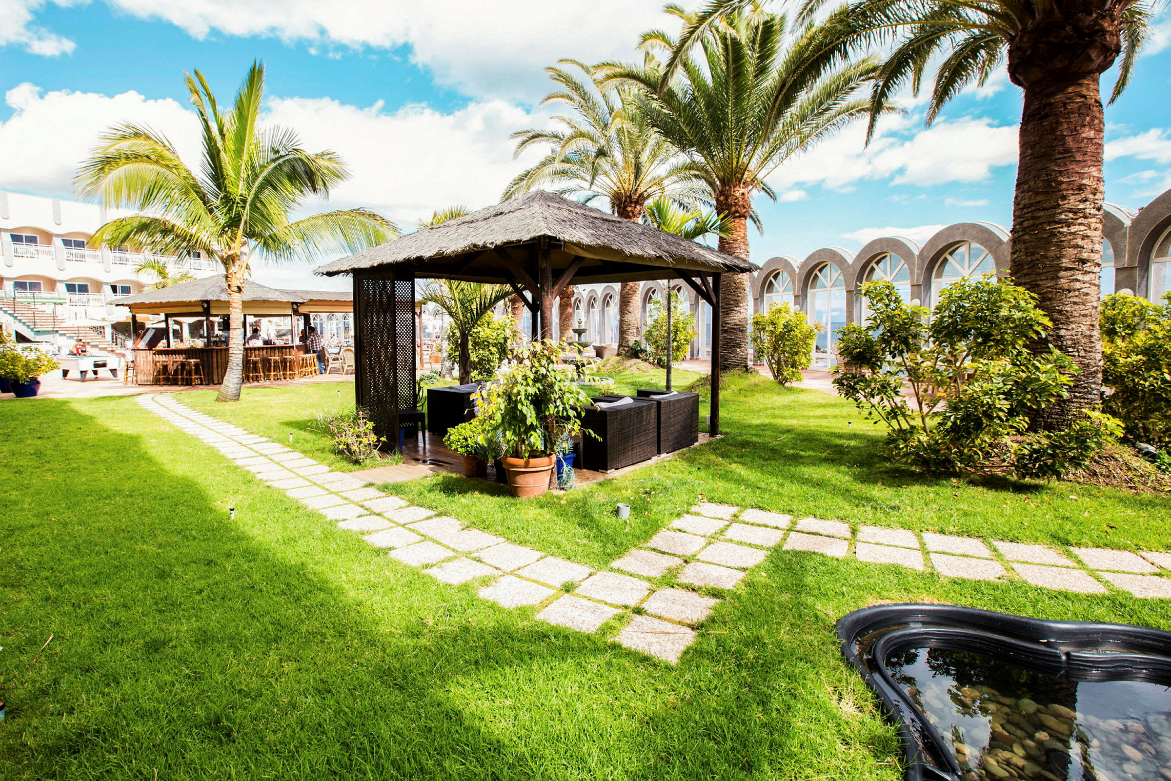 En grönskande trädgård med en pergola och möblerad sittgrupp, omgiven av palmer och buskar. (AI-genererad text)