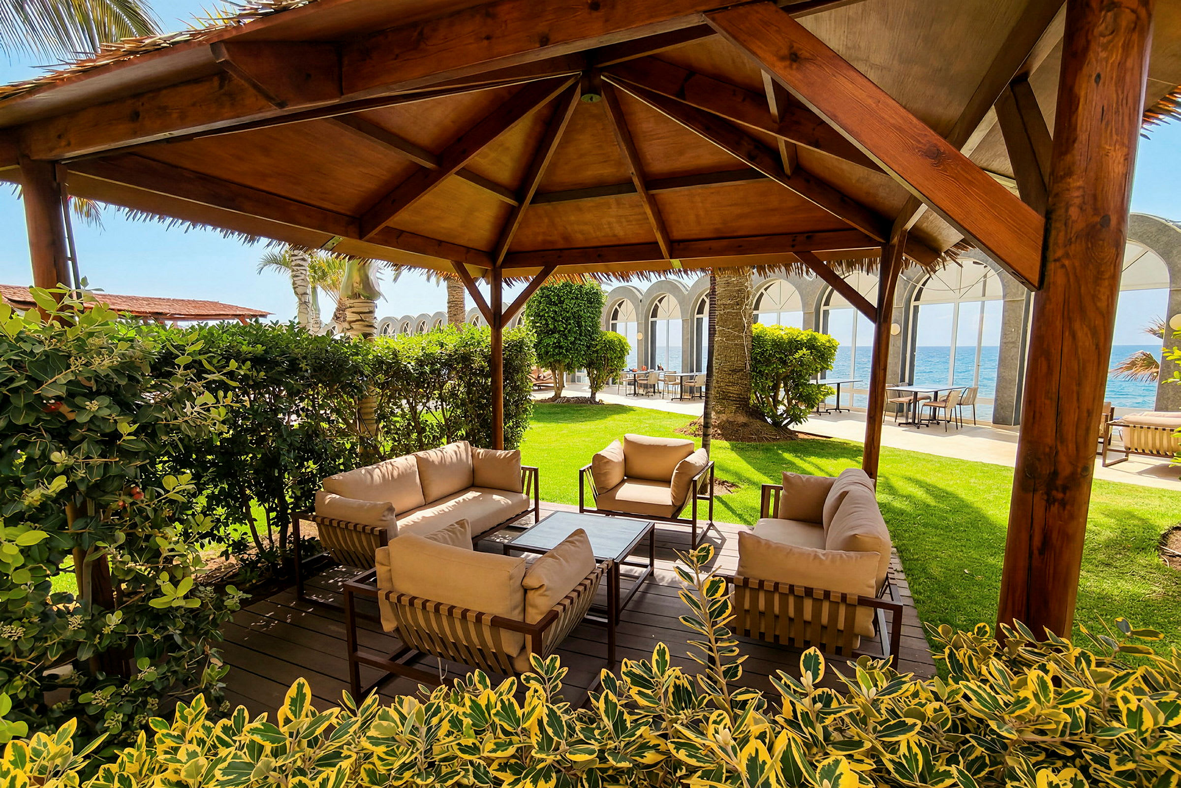 En sittgrupp med soffor och stolar under en träpergola, omgiven av grönska. I bakgrunden finns havet och en byggnad med bågformade fönster. (AI-genererad text)