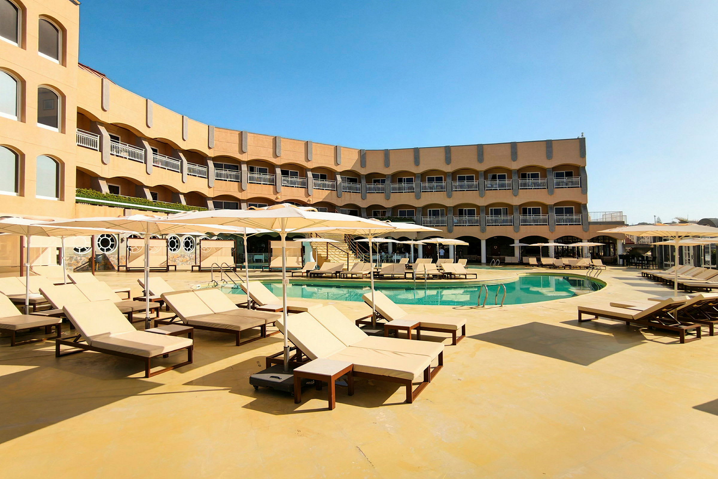 En hotellpool med solstolar och parasoller runt om. Hotellets byggnad syns i bakgrunden. (AI-genererad text)